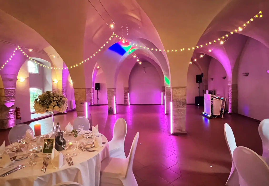 Festlich beleuchtete Eventlocation mit dekorierten Rundbogen und lila Lichtakzenten – Blick auf festlich gedeckten Tisch und DJ-Pult im Hintergrund.