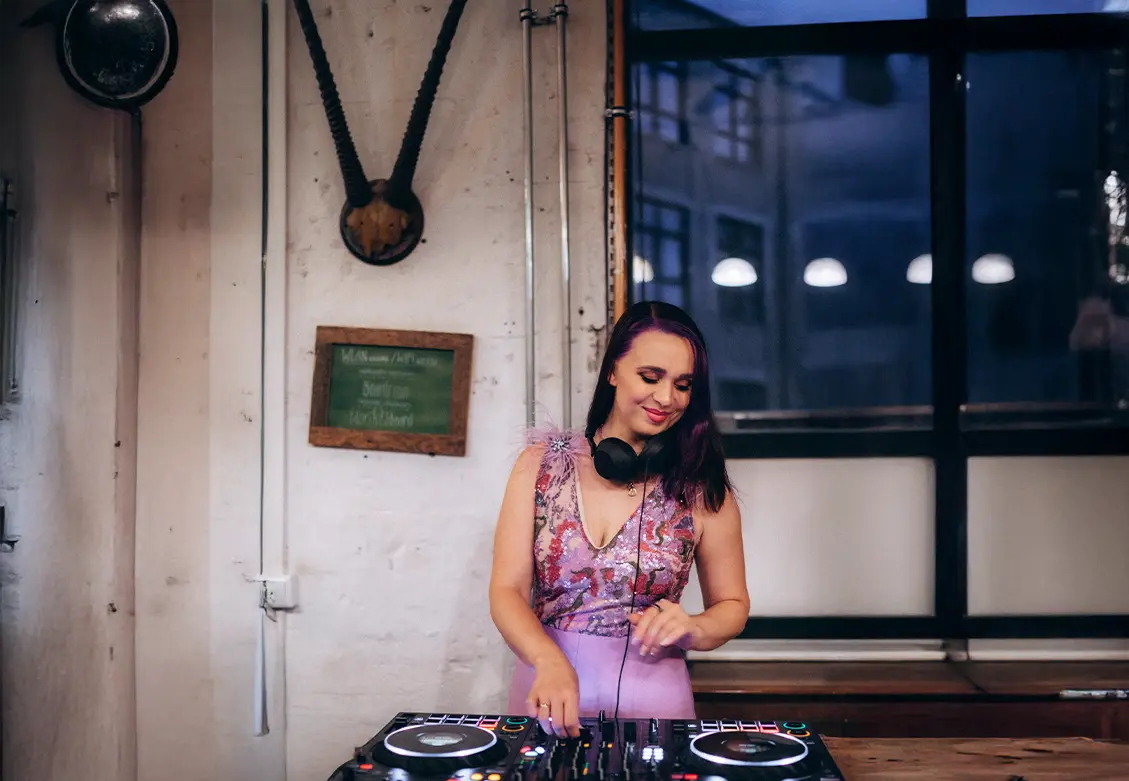 DJane Katrin Mendelsohn legt auf einem stilvollen Event mit violettem Glitzerkleid auf – konzentriert und strahlend hinter dem DJ-Pult.