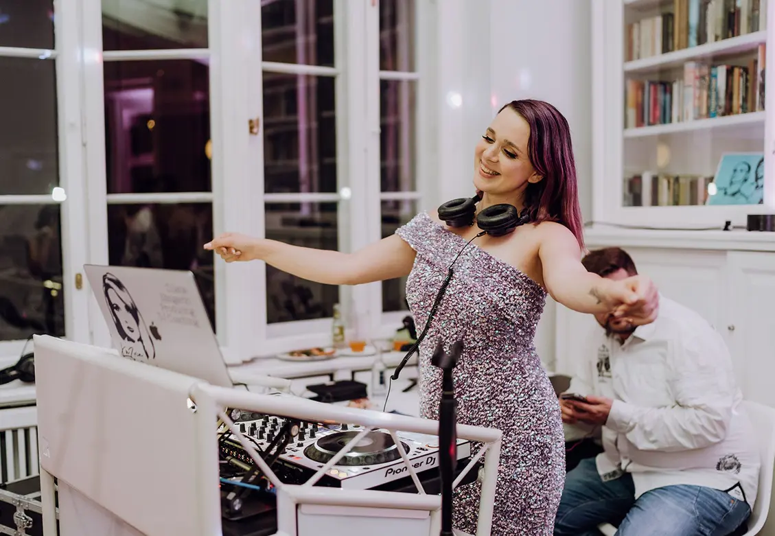 DJane Katrin Mendelsohn strahlt hinter ihrem weißen DJ-Pult mit Kopfhörern um den Hals – elegante Atmosphäre in heller Eventlocation.