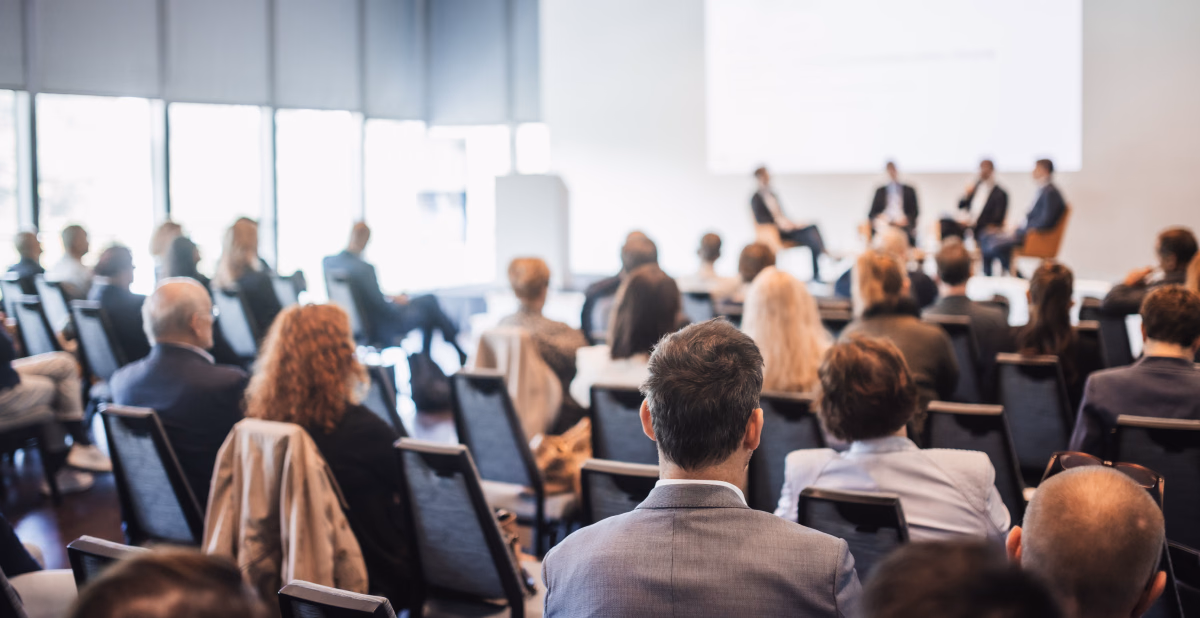 A large audience sits in rows of chairs listening to a panel of speakers on a stage at a conference.