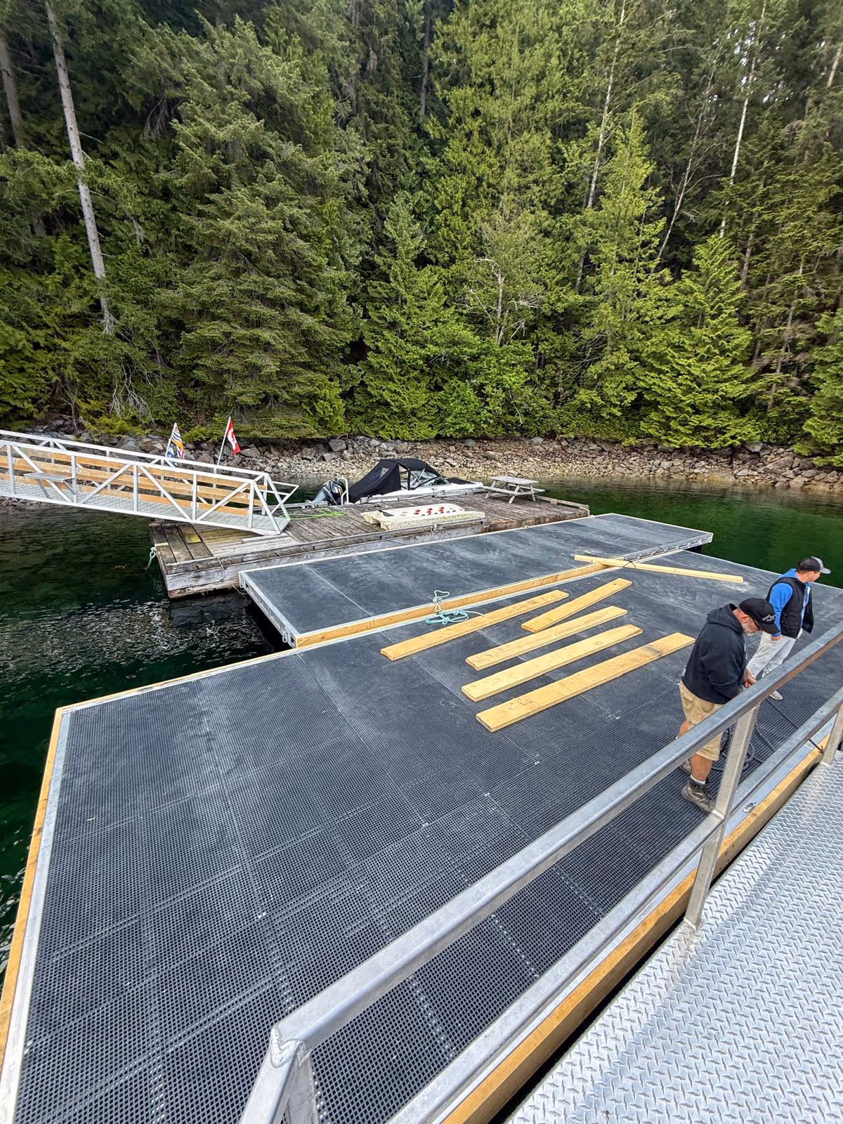 CoastMetalWorks crew installing a large custom aluminum floating dock with non-slip grated decking beside an existing dock and gangway on a remote forested BC inlet
