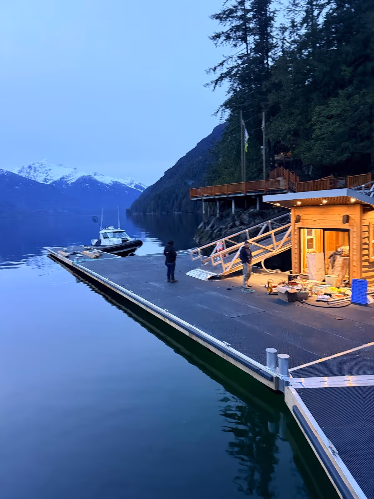 Large custom aluminum floating dock with a lit cedar boathouse and gangway at dusk, moored boat alongside, on a calm snow-capped BC fjord during final installation