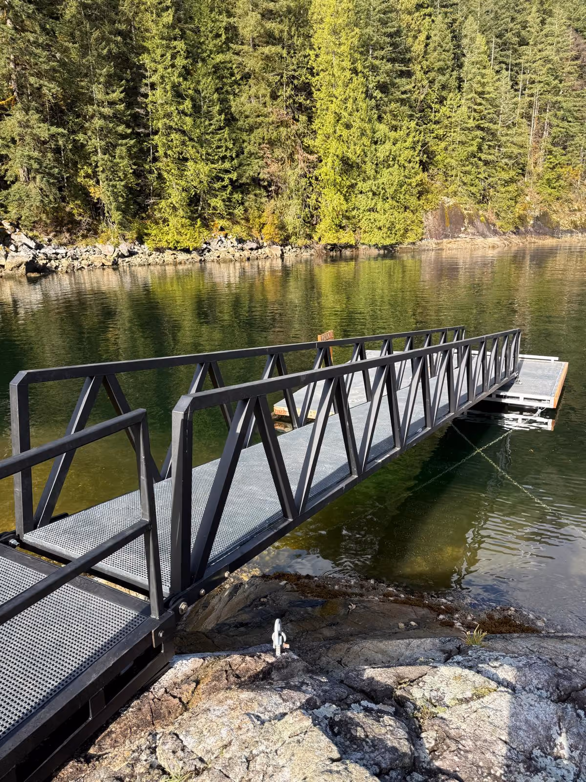 Gunmetal powder-coated aluminum gangway ramp with truss-style railings spanning from a rocky shoreline to a custom floating dock on a forested BC inlet
