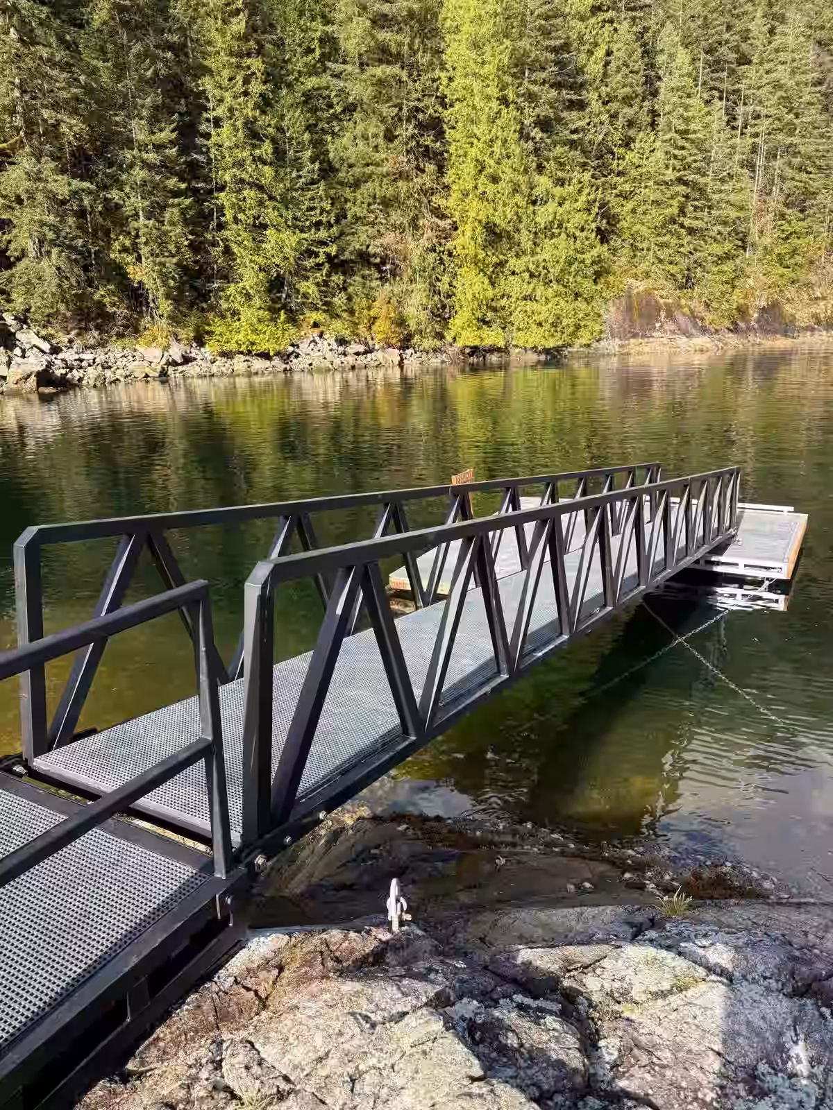 Gunmetal powder-coated aluminum gangway ramp with truss-style railings spanning from a rocky shoreline to a custom floating dock on a forested BC inlet