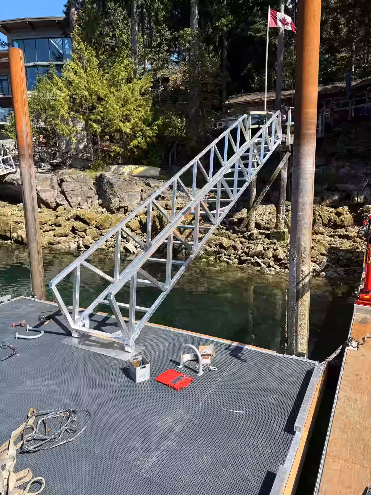 Custom aluminum truss gangway being installed, spanning from a piling-supported floating dock up to a waterfront home's shore access on a rocky BC coastline