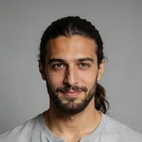 Portrait of a man with dark hair tied back, beard, and mustache, wearing a light gray shirt against a gray background.