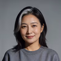 Portrait of a smiling woman with black hair and a gray streak, wearing a gray sweatshirt against a gray background.