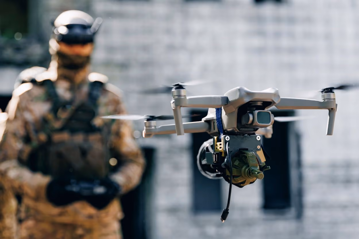 Military drone flying with a mounted device, soldier in camouflage holding controller in the background.