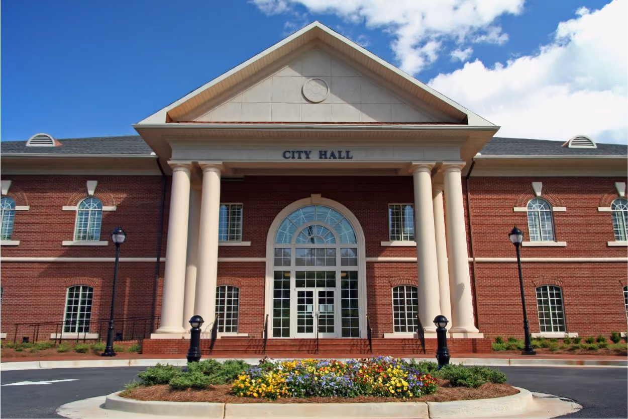Front view of a red brick city hall with tall white columns and a flower bed in front.