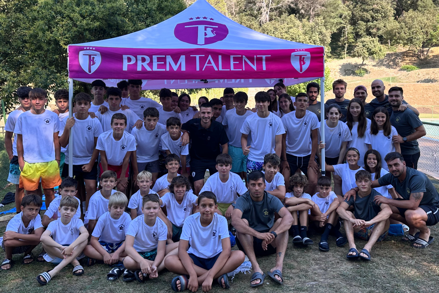 Grup de nens i entrenadors del campus Prem Talent posant junts sota una carpa amb el logotip de l’esdeveniment.