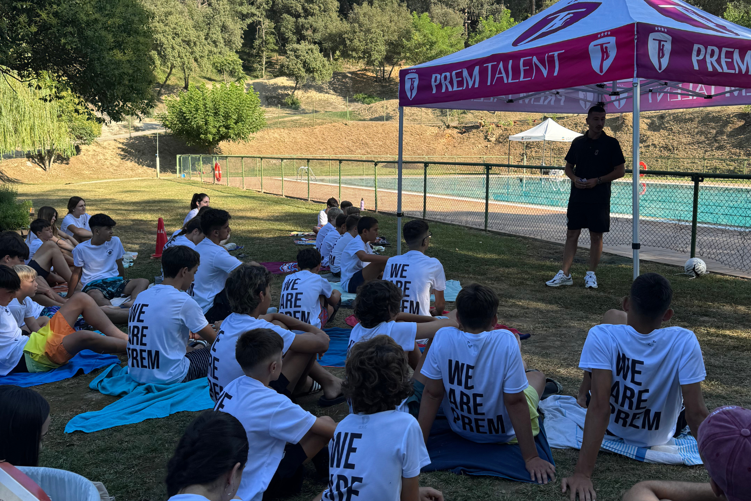 Entrenador del Prem Talent parlant als participants asseguts a l’aire lliure sota la carpa del campus, al costat de la piscina.
