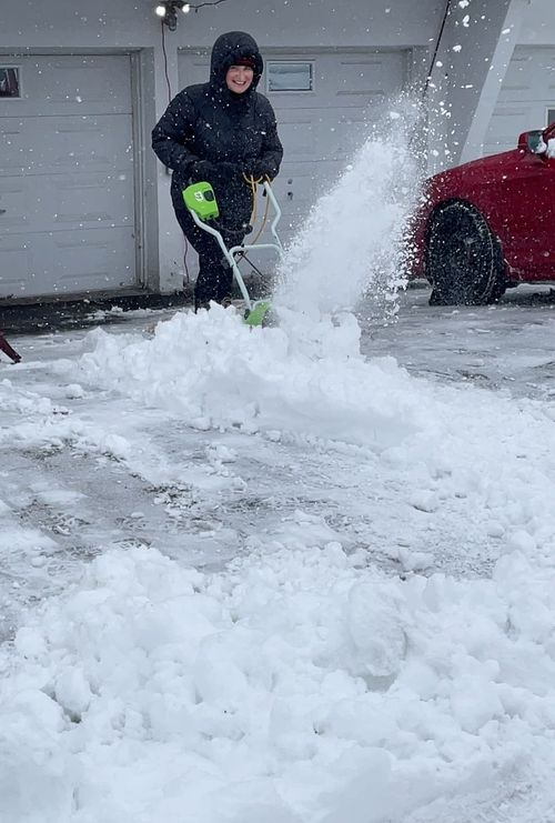 Image of Suzanne in a black jacket and hood smiling while shoveling snow on a driveway with an electric snow shovel with snow spraying in the air.