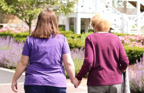 A caregiver and a senior resident walking hand in hand through a garden.