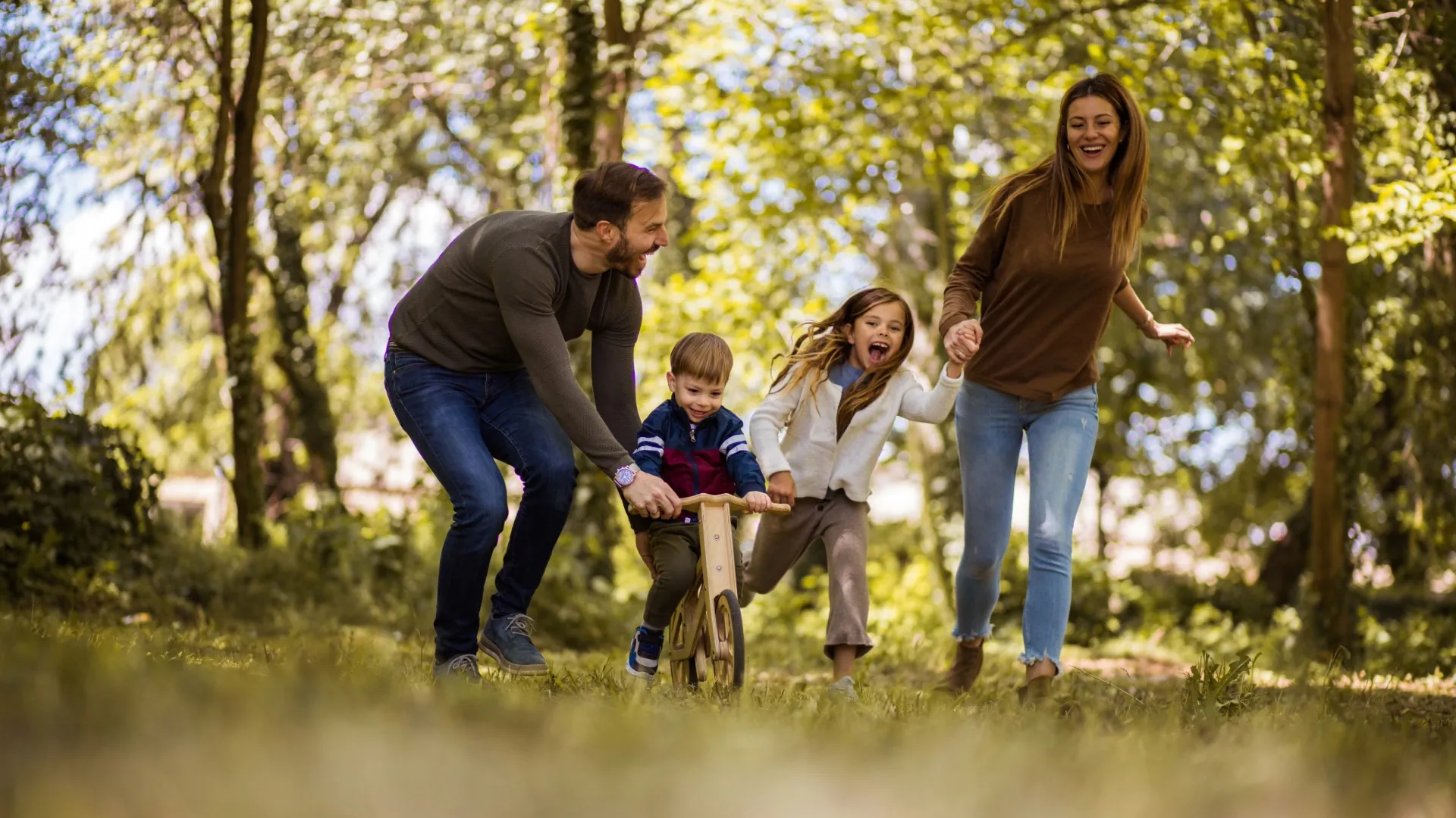 Mom and dad helping their two kids ride bikes along a sunny wooded park path