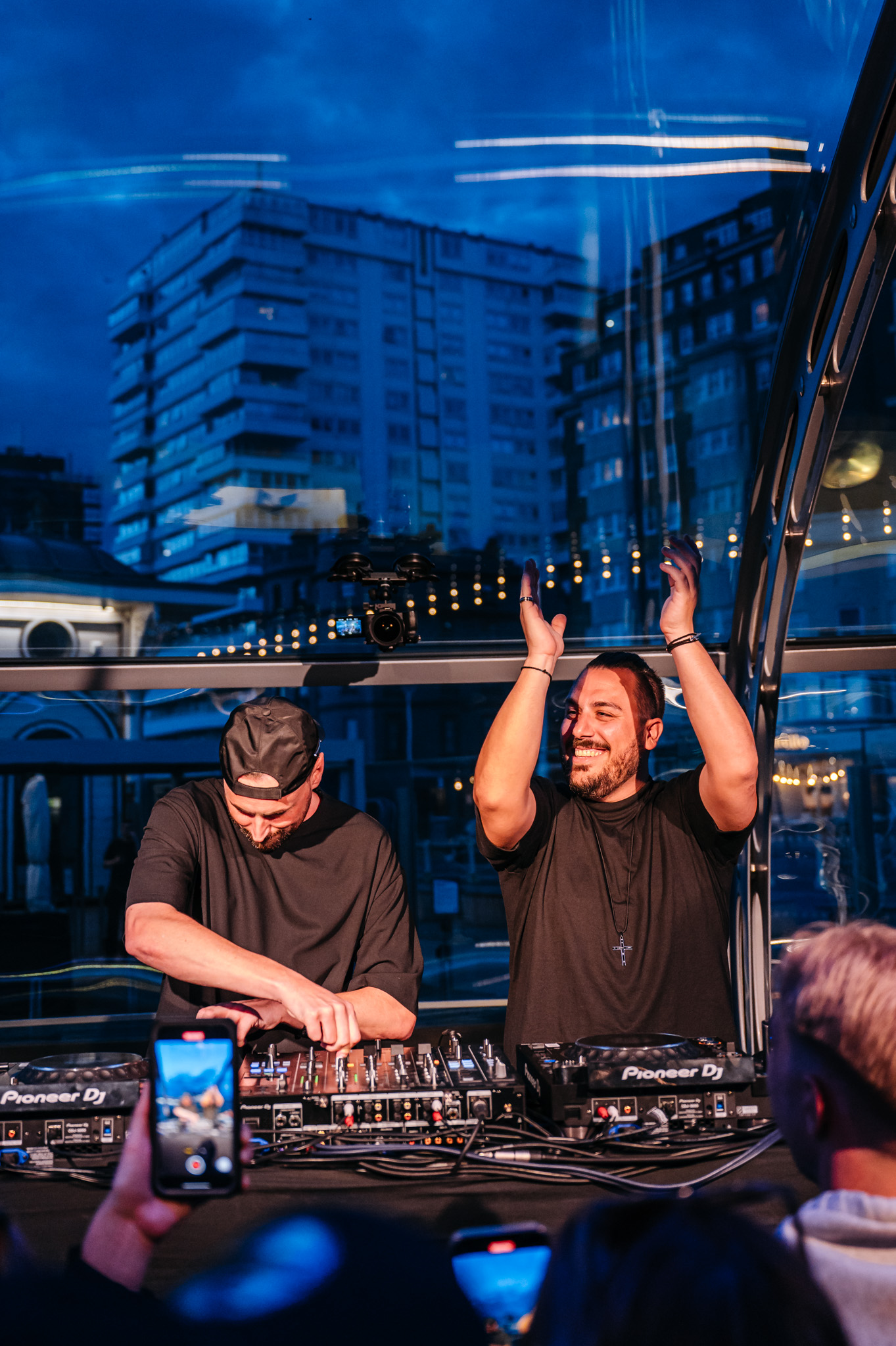 Two DJs perform with Pioneer DJ equipment at an outdoor nighttime event with city buildings in the background.