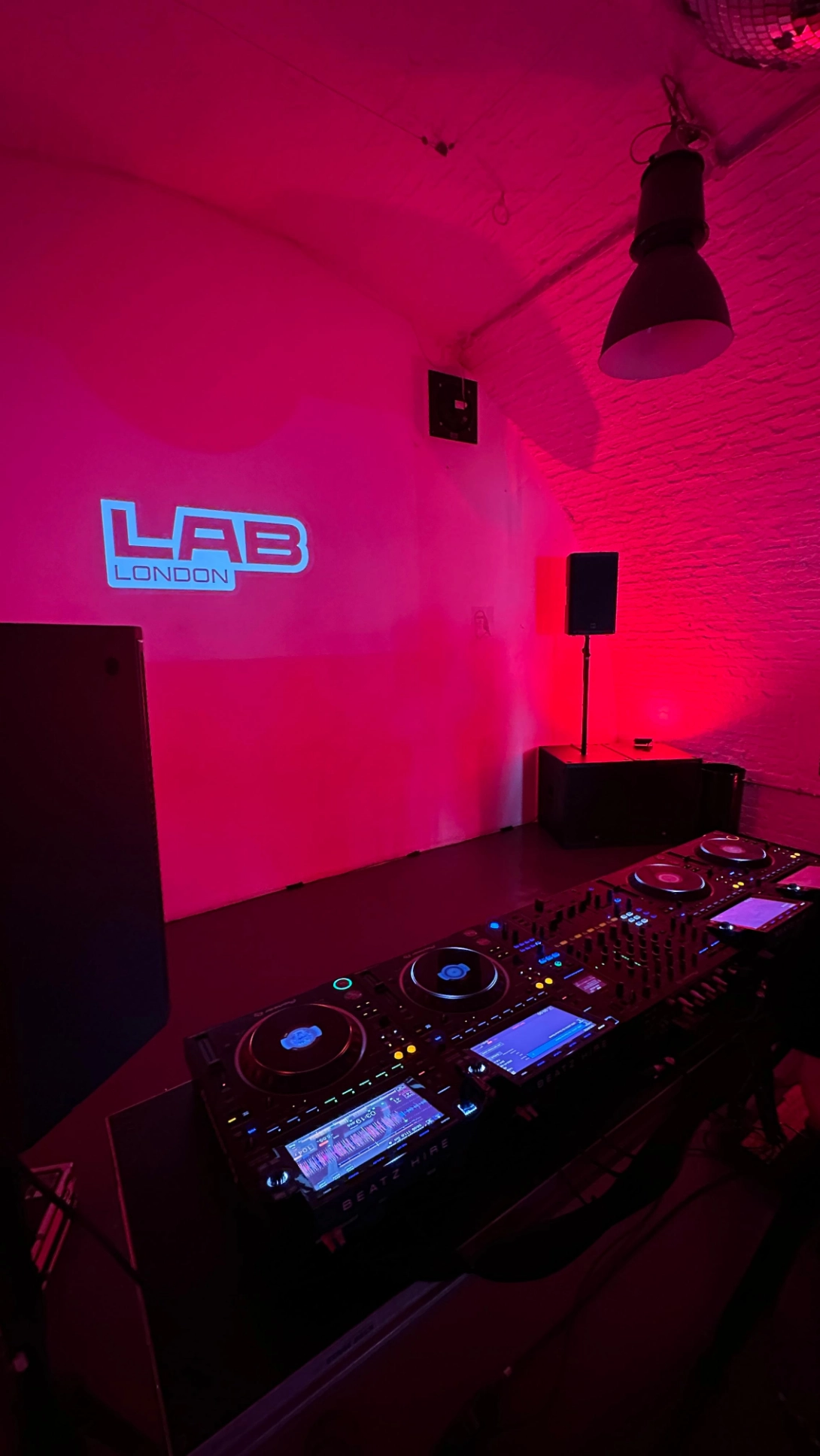 DJ mixing console with illuminated screens and controls in a dimly lit room with pink and red lighting and a LAB London logo projected on the wall.