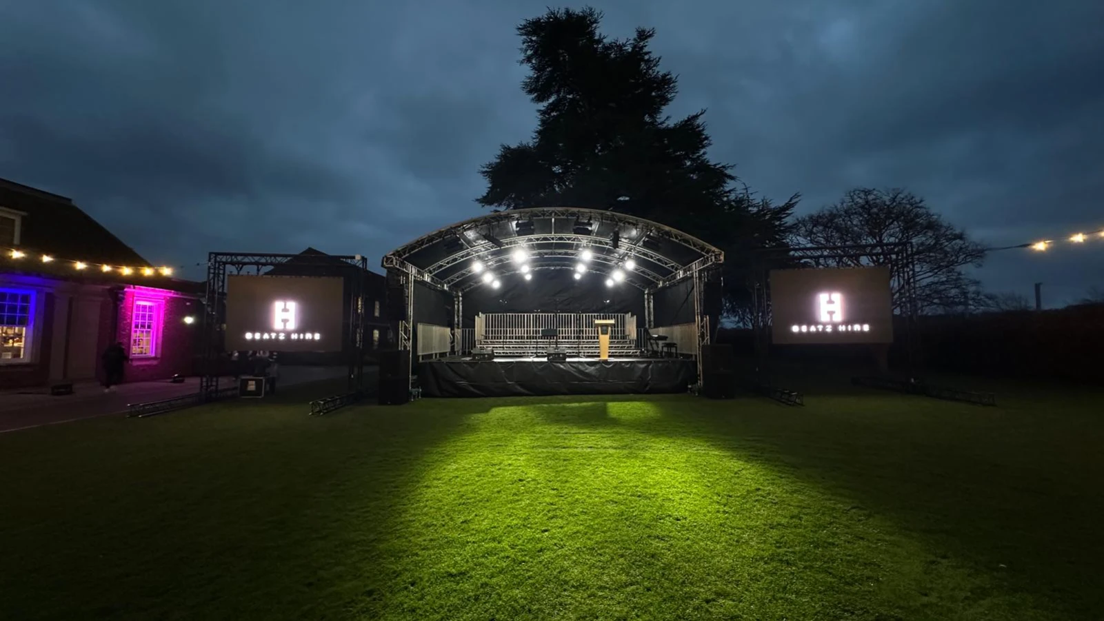 Outdoor stage at night with overhead lights, podium in the center, and two large screens displaying 'Beatz Hire' on either side.