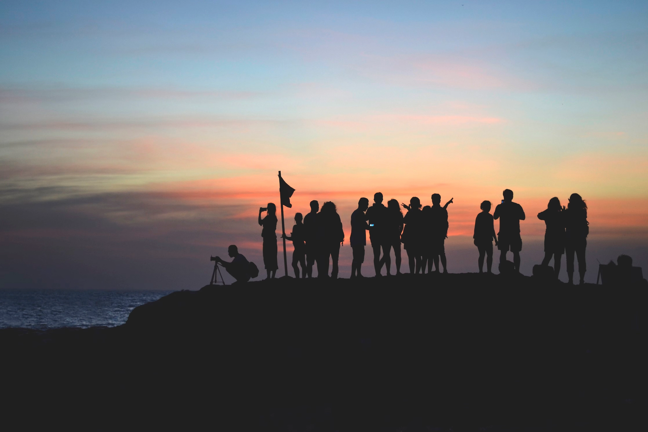 Silhouettes of a group of people on a hill by the ocean at sunset with a colorful sky.