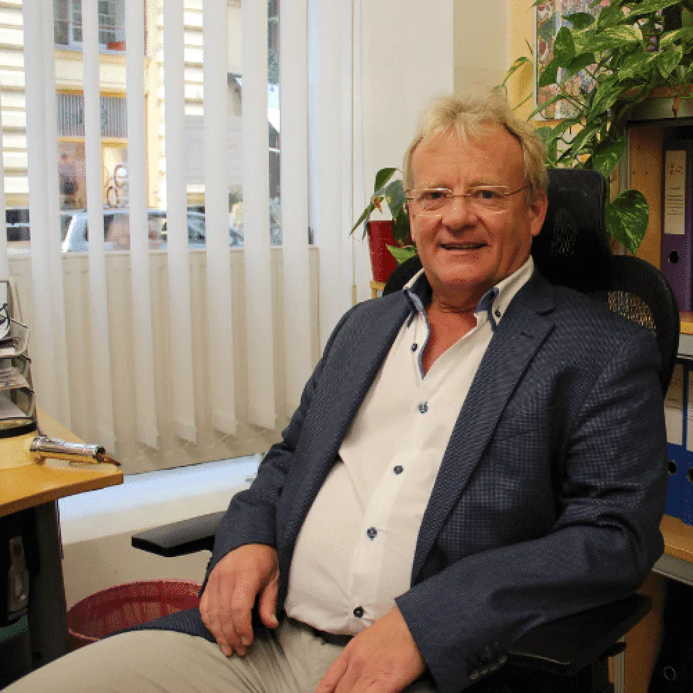 Ein Mann in einem blauen Anzug und weissem Hemd sitzt entspannt in einem Bürostuhl. Hinter ihm stehen Pflanzen, Regale mit Ordnern und ein Fenster mit vertikalen Jalousien. Die Szene vermittelt ein professionelles und einladendes Arbeitsumfeld, passend zur Qualität und Zuverlässigkeit von Nextlevel Handwerk, der Werbeagentur aus Schlanders, Meran, Südtirol.  