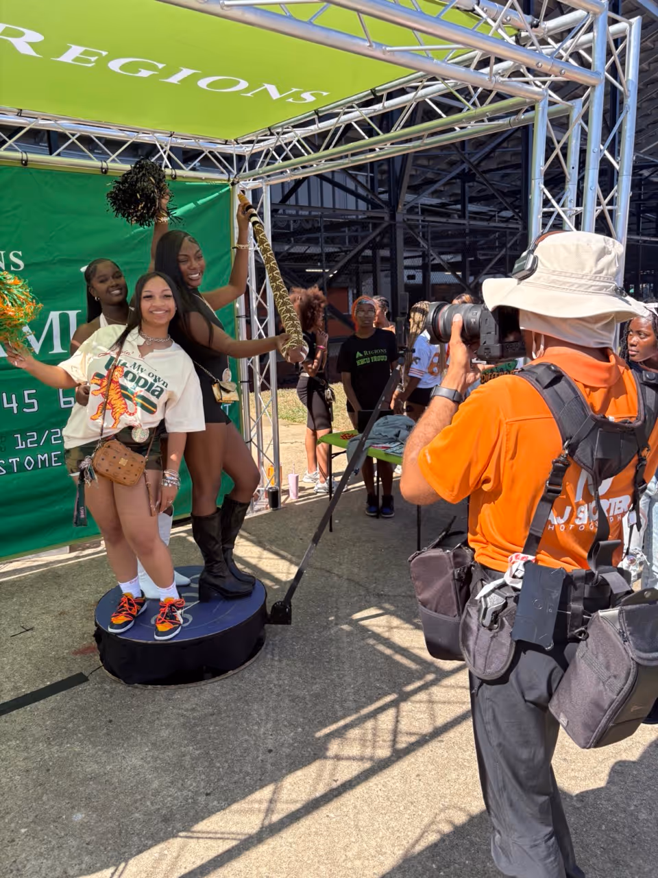 Three young women posing with pom-poms and a megaphone on a small round platform under a green canopy while a photographer in an orange shirt takes their picture.