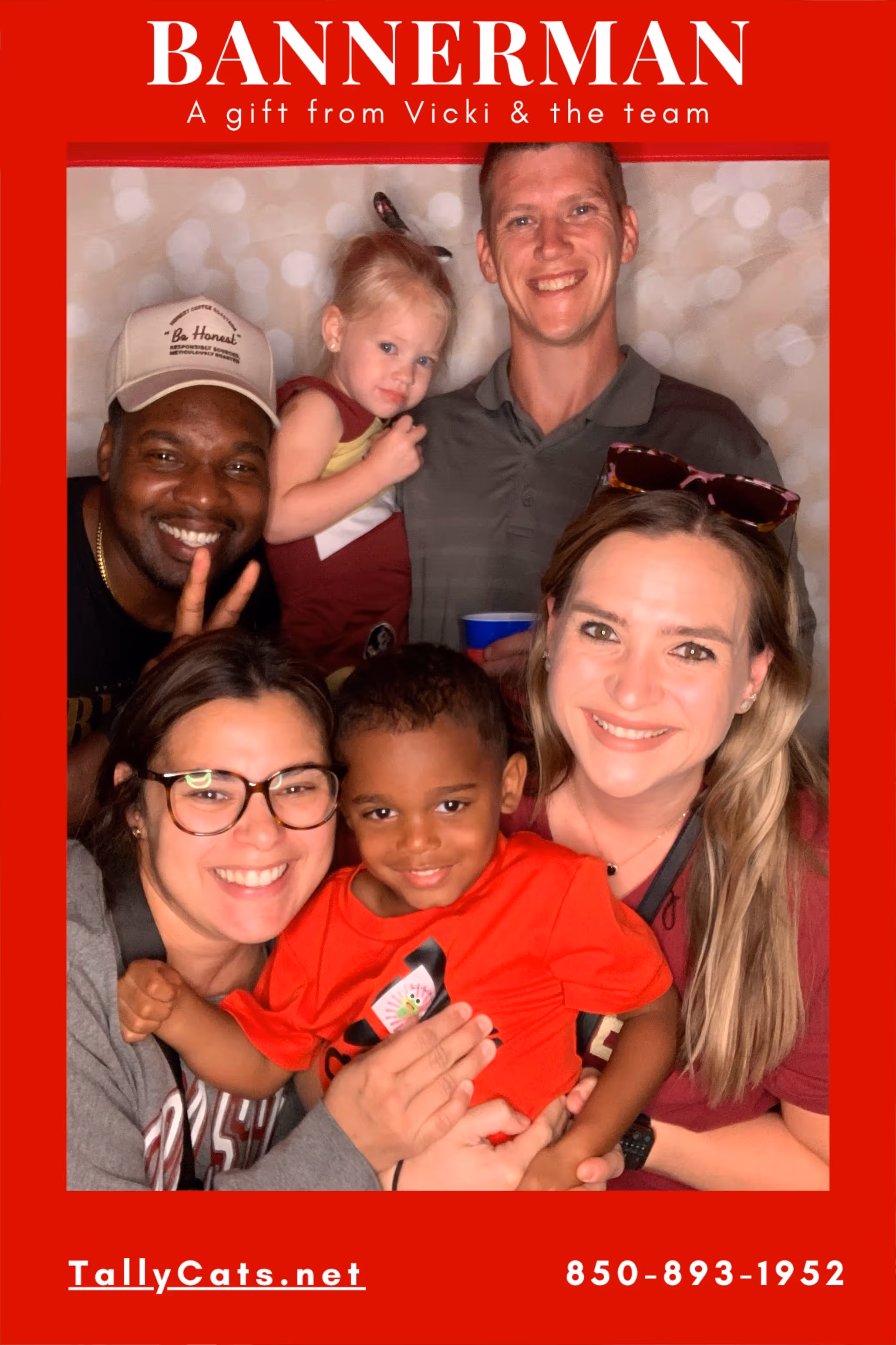 Six smiling people, including two children, posing closely together in a red-framed photo booth picture with the text 'BANNERMAN A gift from Vicki & the team' at the top and 'TallyCats.net 850-893-1952' at the bottom.