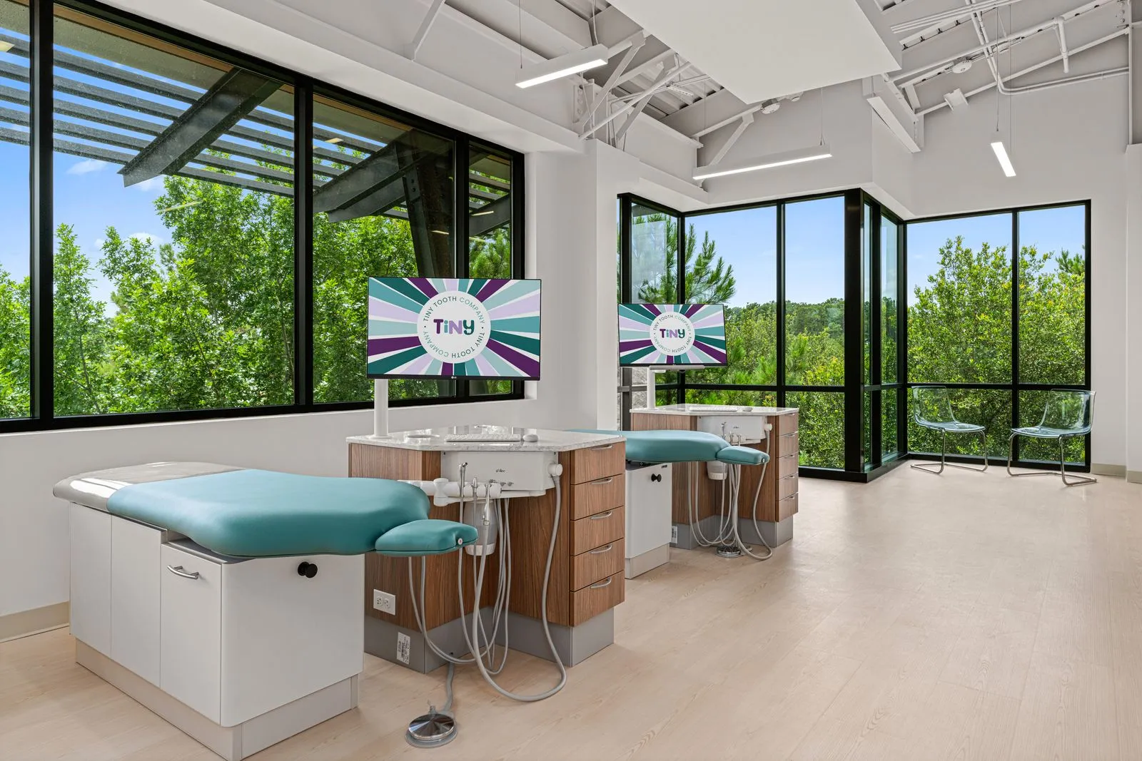 Open bay treatment area at Tiny Tooth Company with two dental chairs, mounted monitors displaying the clinic logo, and panoramic views of lush greenery through large floor-to-ceiling windows.