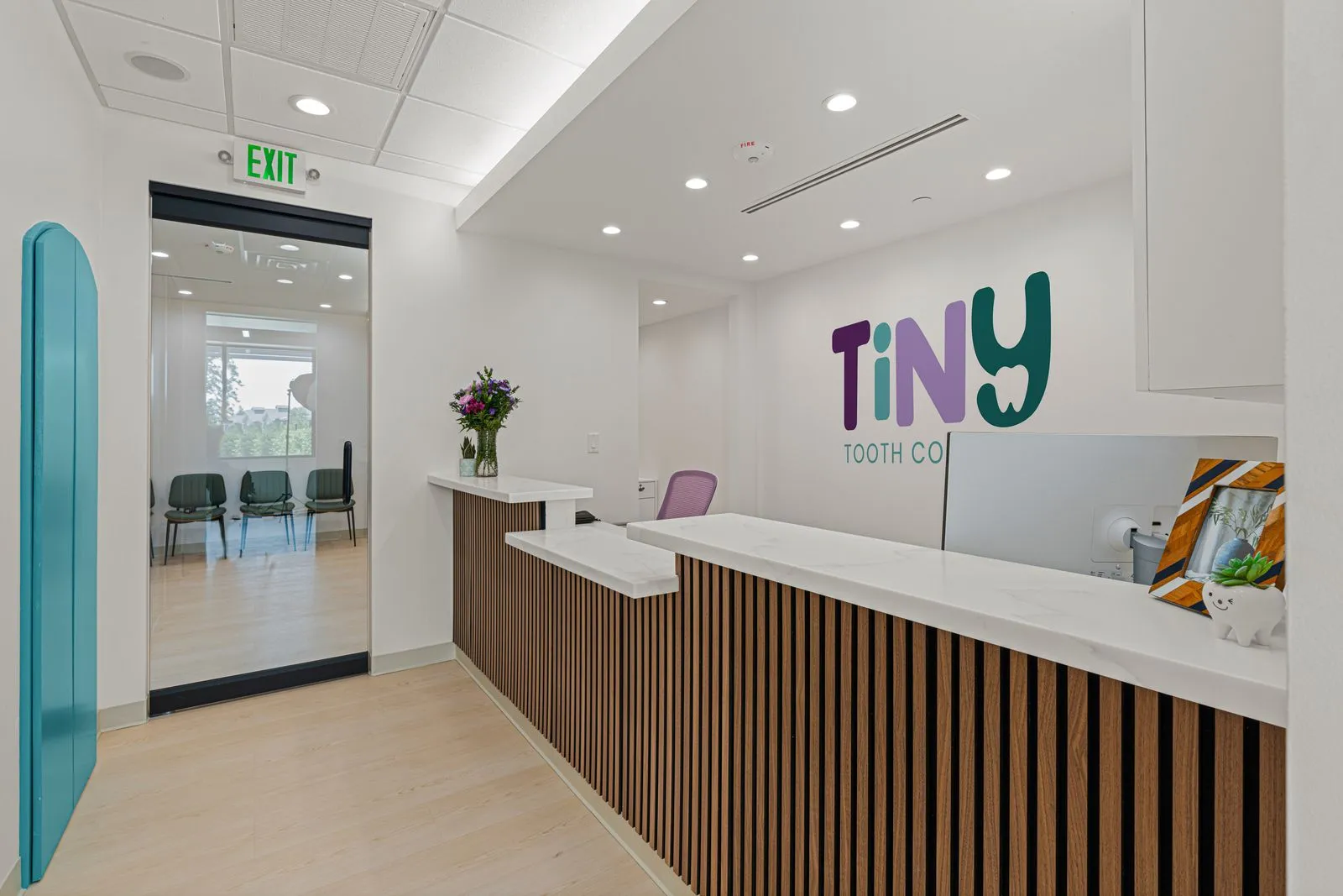 Reception area at Tiny Tooth Company with a wood-accented marble desk, fresh flowers, and the clinic’s colorful logo on the wall, with a view into the adjacent waiting room.