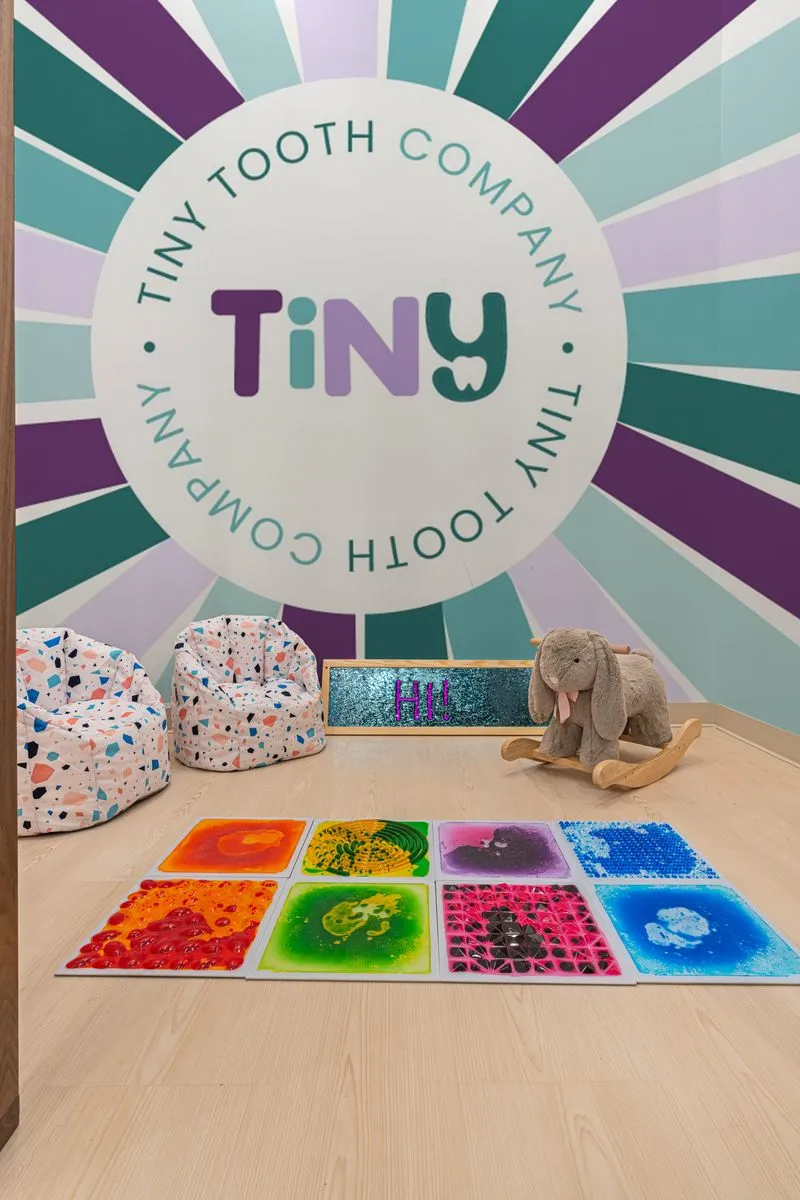 Colorful kids’ play area at Tiny Tooth Company, featuring sensory floor tiles, terrazzo-print bean bags, a plush rocking bunny, and a vibrant mural with the clinic’s logo.