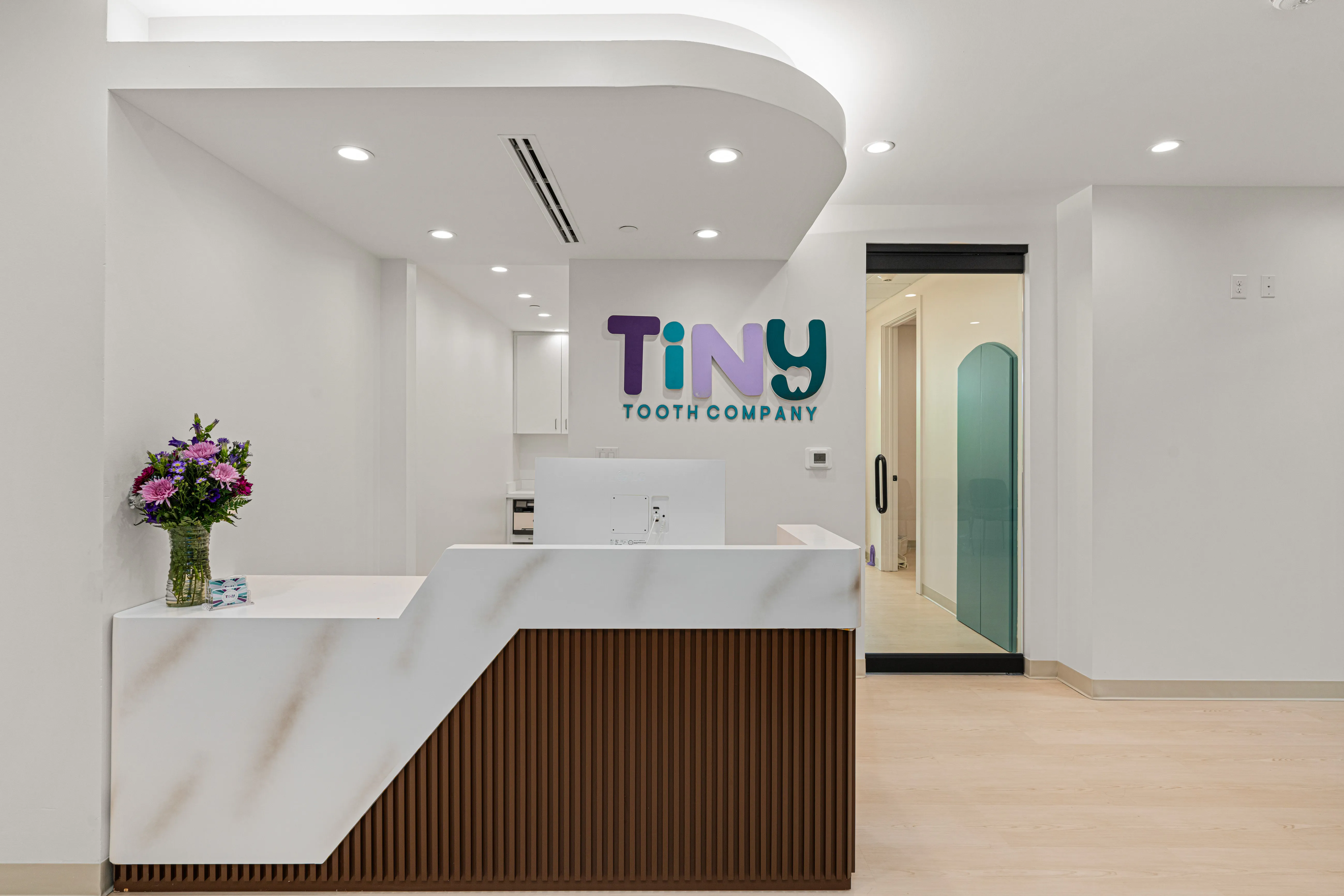 Reception area at Tiny Tooth Company pediatric dental clinic, featuring a modern white marble front desk, fresh flowers, and colorful logo signage on the wall.