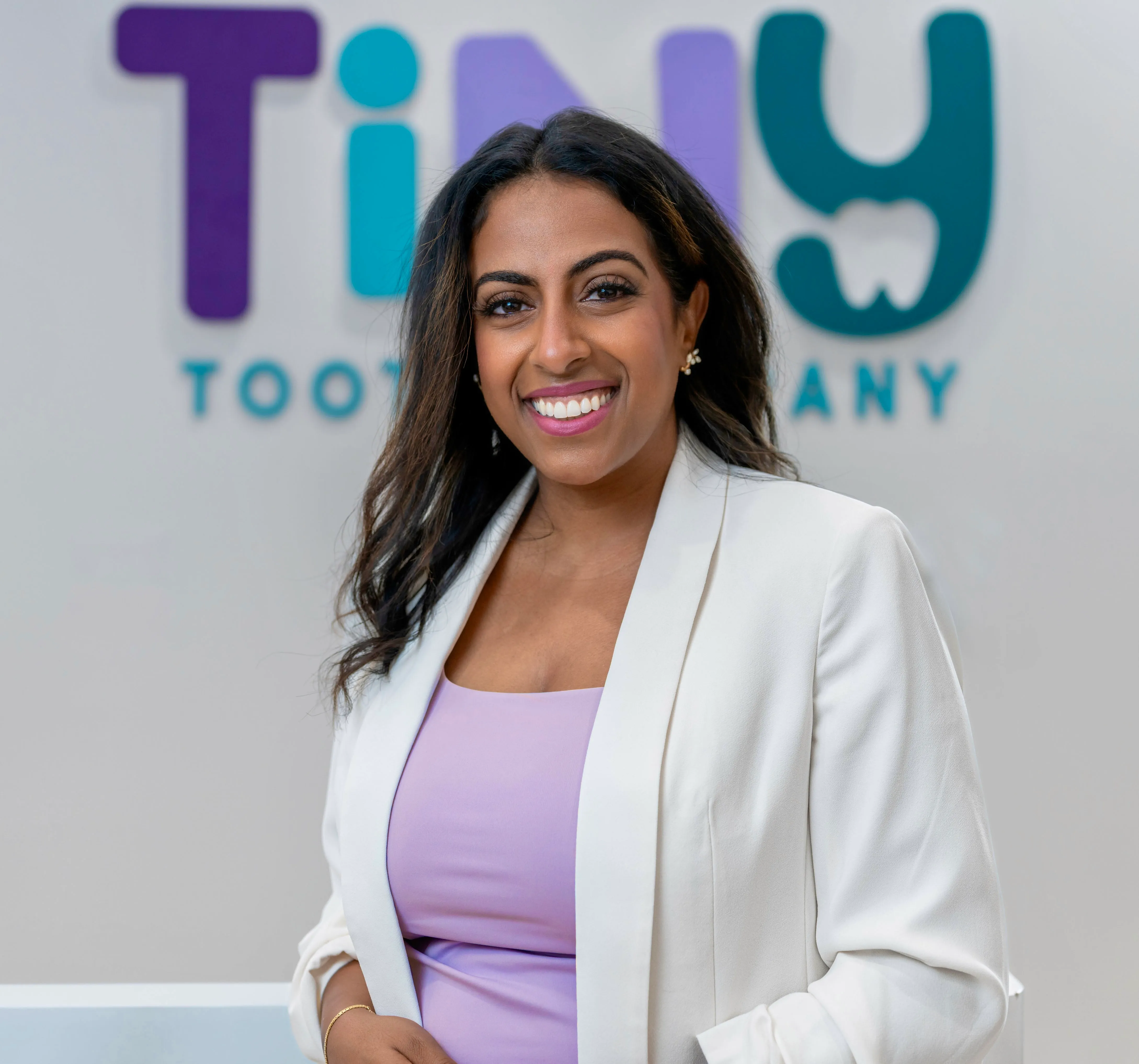 Dr. Sarah Arafat, founder of Tiny Tooth Company, smiling warmly in a white blazer and lavender dress, standing in front of the pediatric dental clinic’s logo wall.