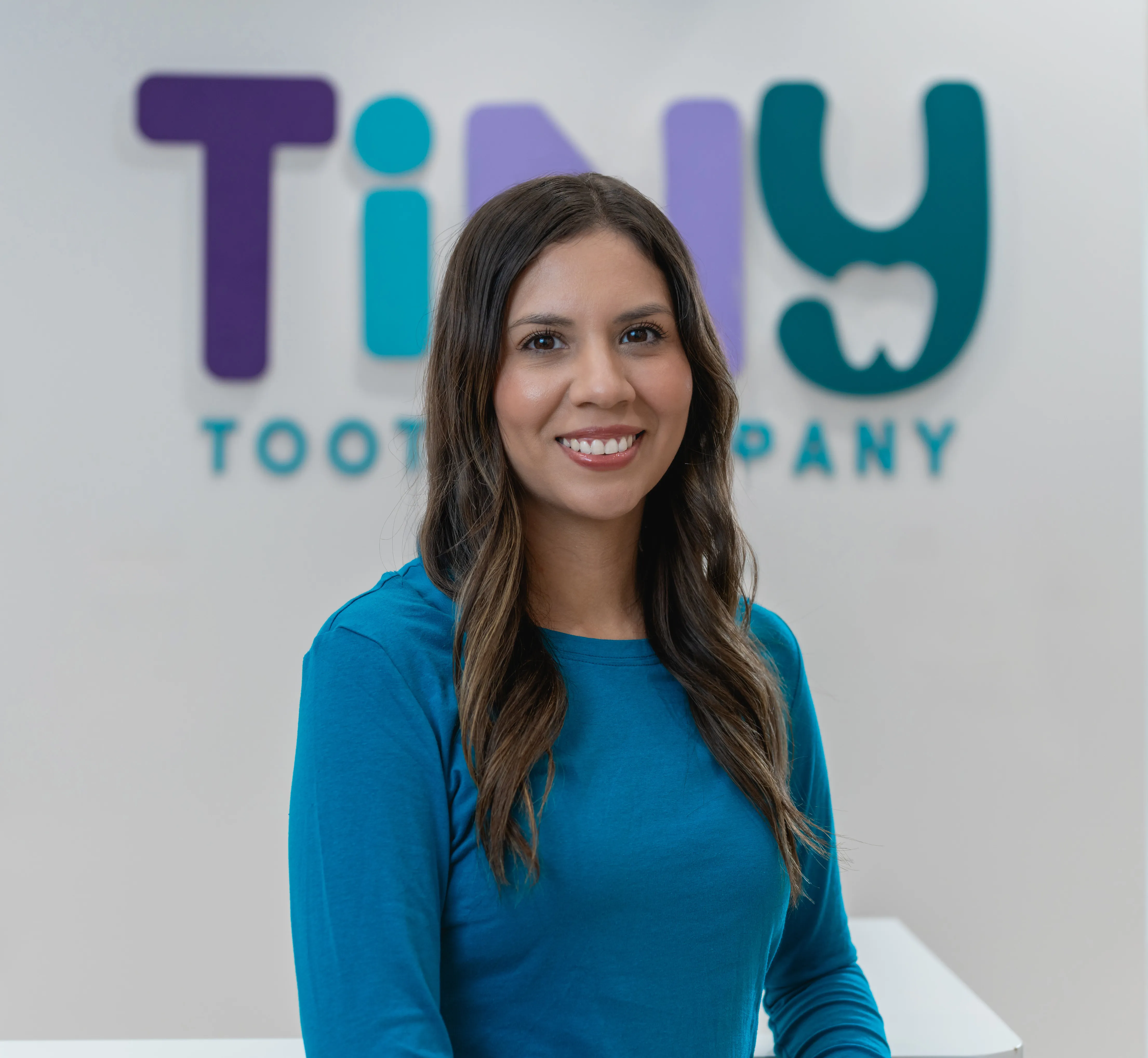 Team member at Tiny Tooth Company smiling confidently in a teal uniform, posed in front of the clinic’s colorful logo wall.