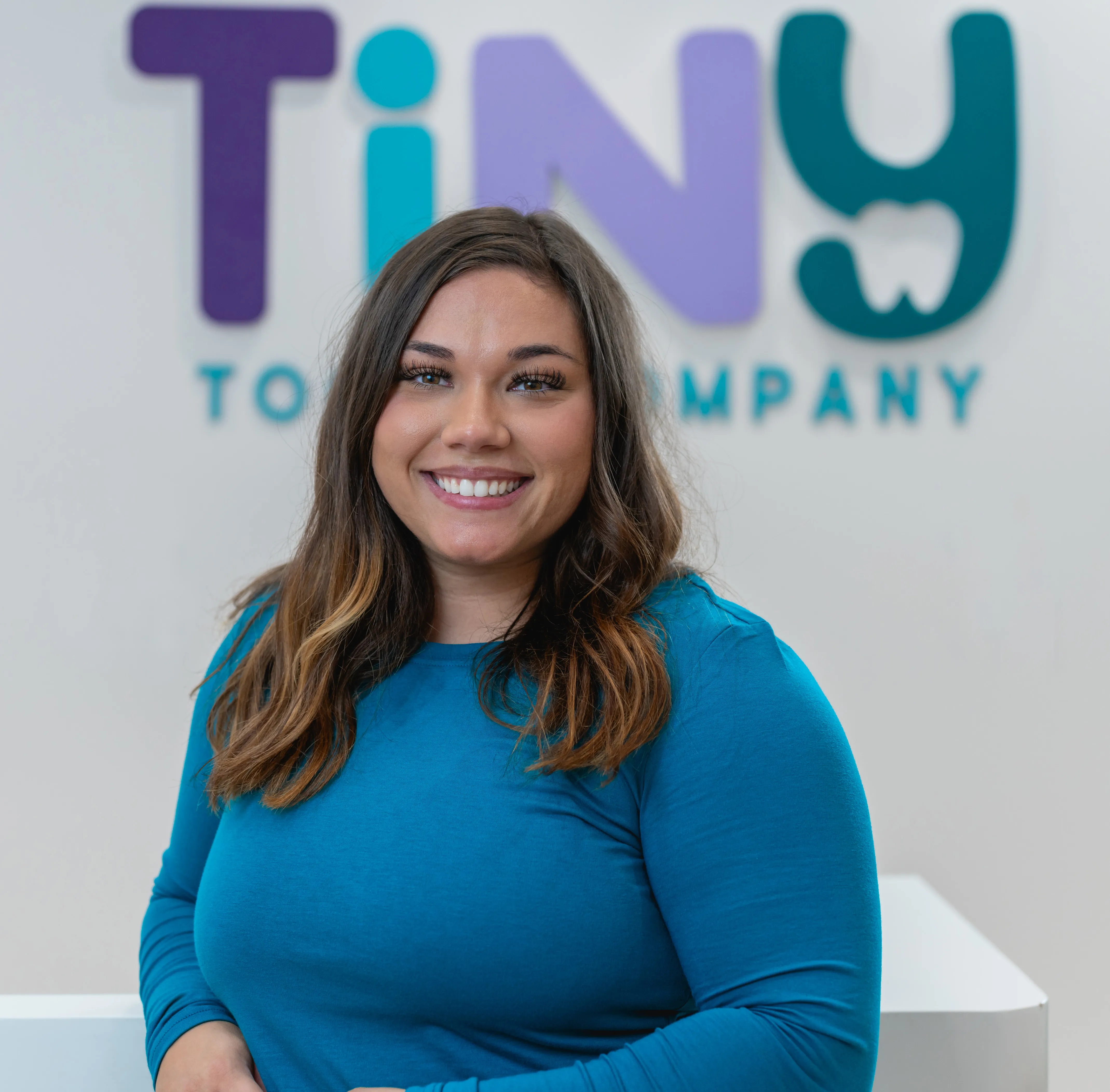 Smiling team member at Tiny Tooth Company in a teal uniform, posed confidently in front of the clinic’s colorful logo wall.