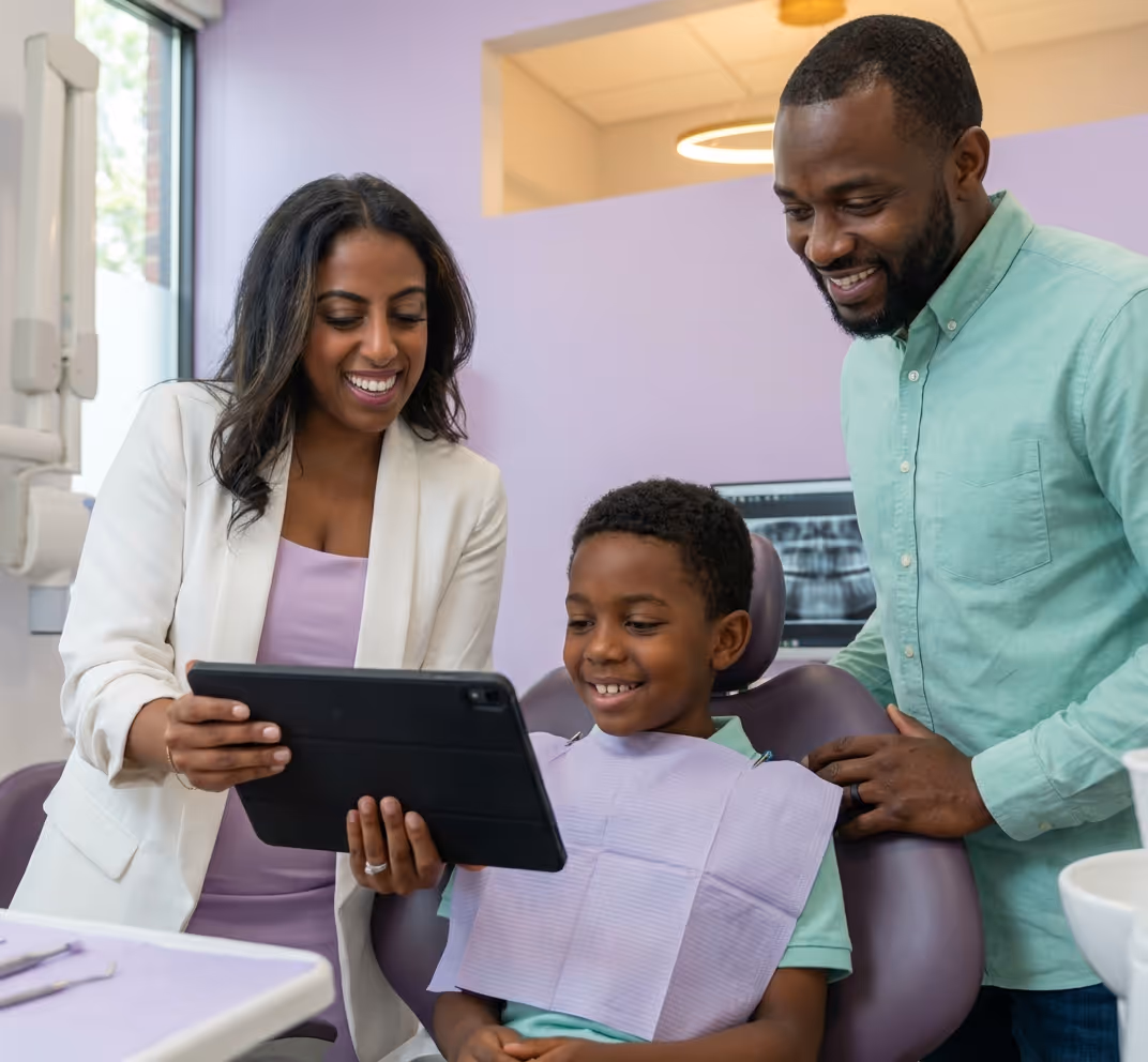 Dentist with family reviewing tablet
Female pediatric dentist with curly dark hair in a white coat reviewing dental care information on a tablet alongside a smiling father in a teal shirt and his young son seated between them, during a family-focused consultation in a bright, softly lit exam room at Tiny Dental Company in The Woodlands, TX.
