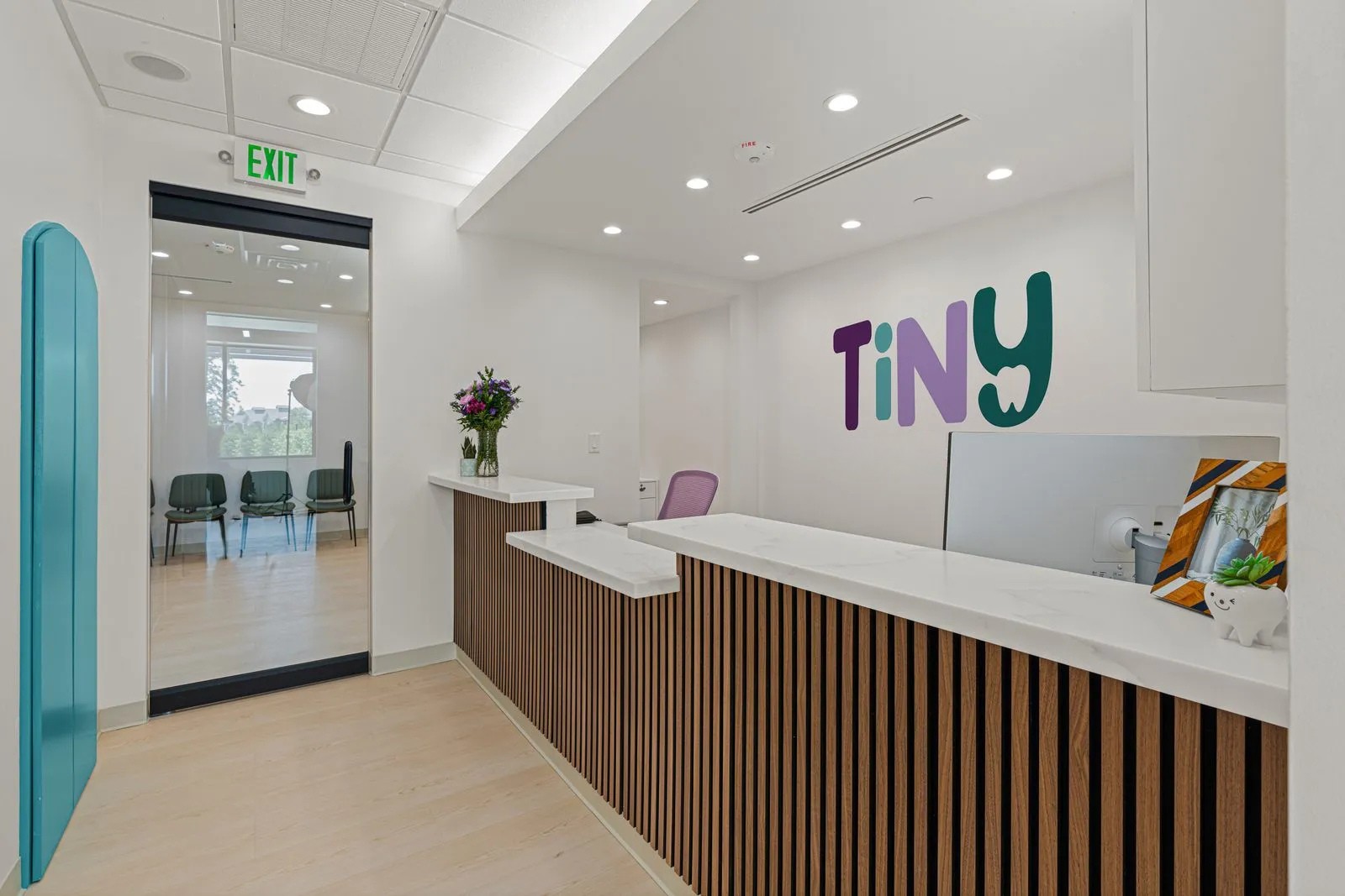Reception area at Tiny Dental Company with a wood-accented marble desk, fresh flowers, and the clinic’s colorful logo on the wall, with a view into the adjacent waiting room.