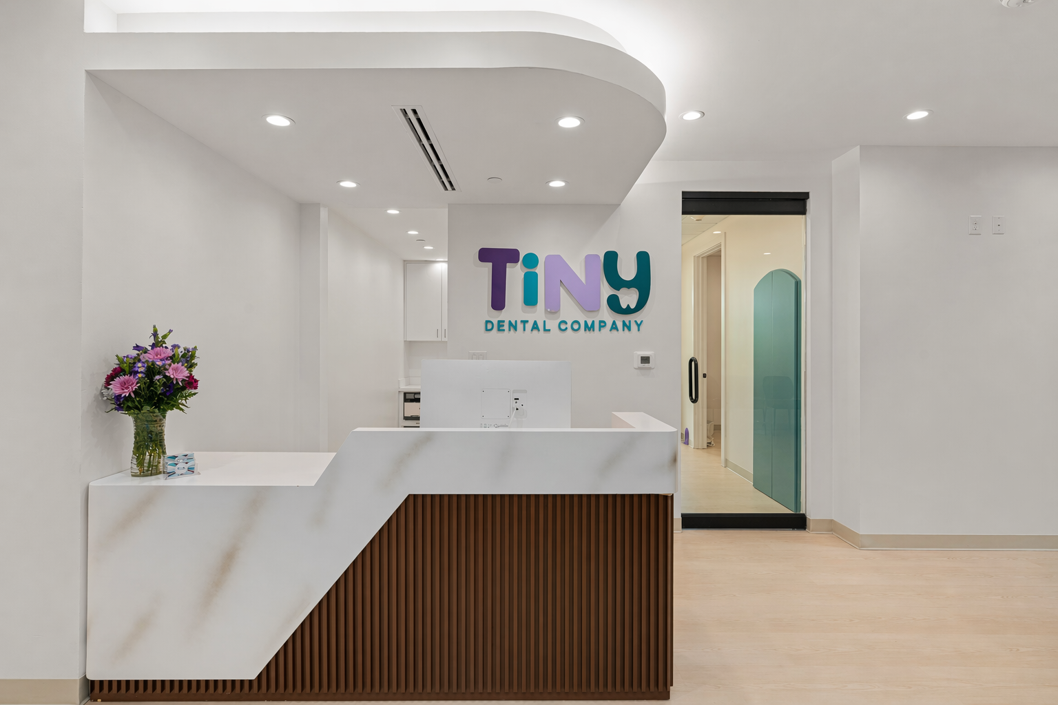 Reception area at Tiny Dental Company pediatric dental clinic, featuring a modern white marble front desk, fresh flowers, and colorful logo signage on the wall.