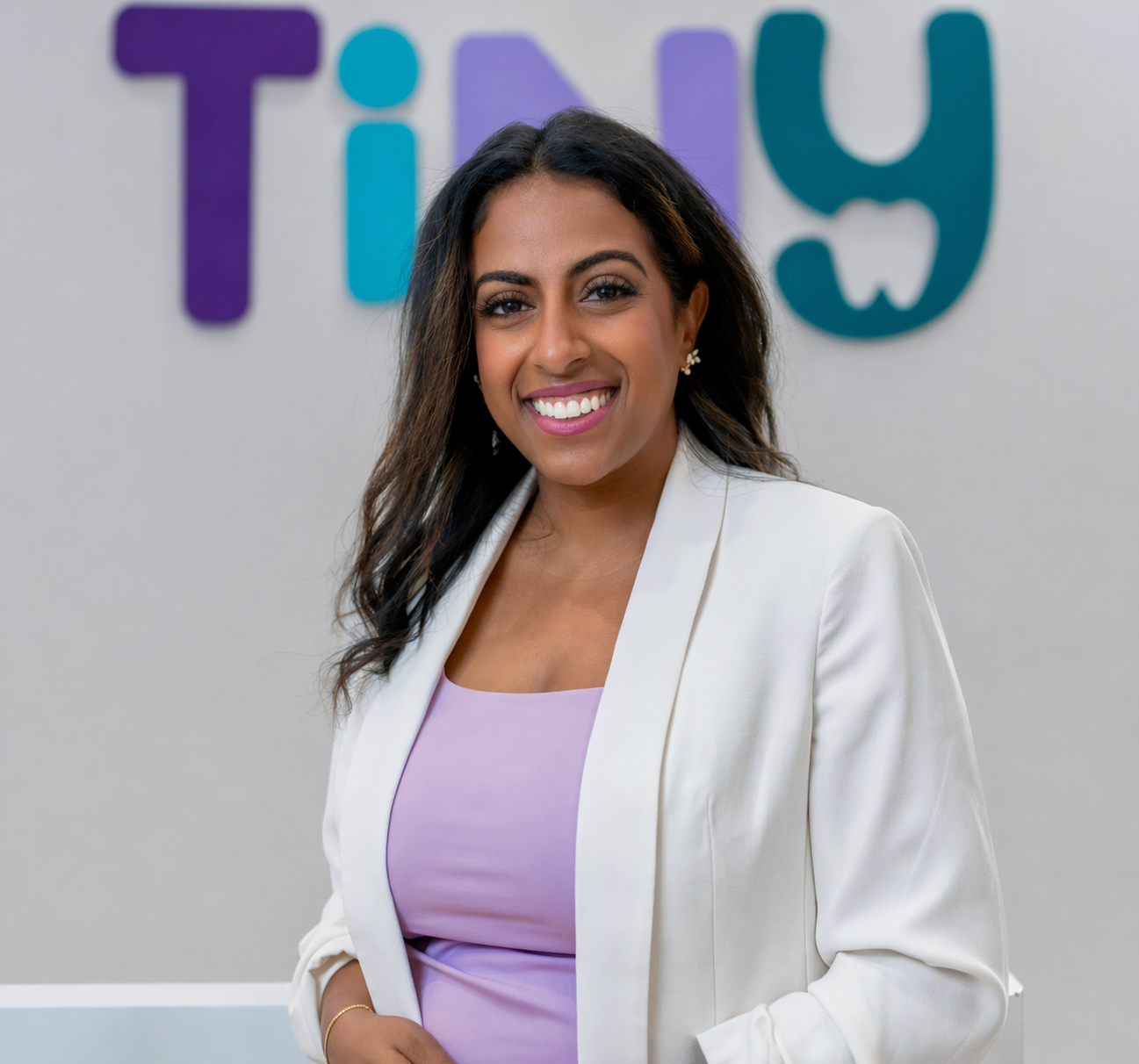 Dr. Sarah Arafat, founder of Tiny Dental Company, smiling warmly in a white blazer and lavender dress, standing in front of the pediatric dental clinic's logo wall.