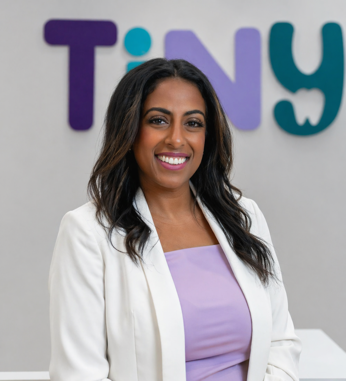 Dr. Sarah Arafat, founder of Tiny Dental Company, smiling in a white blazer and lavender top, posed confidently in front of the clinic’s logo wall.