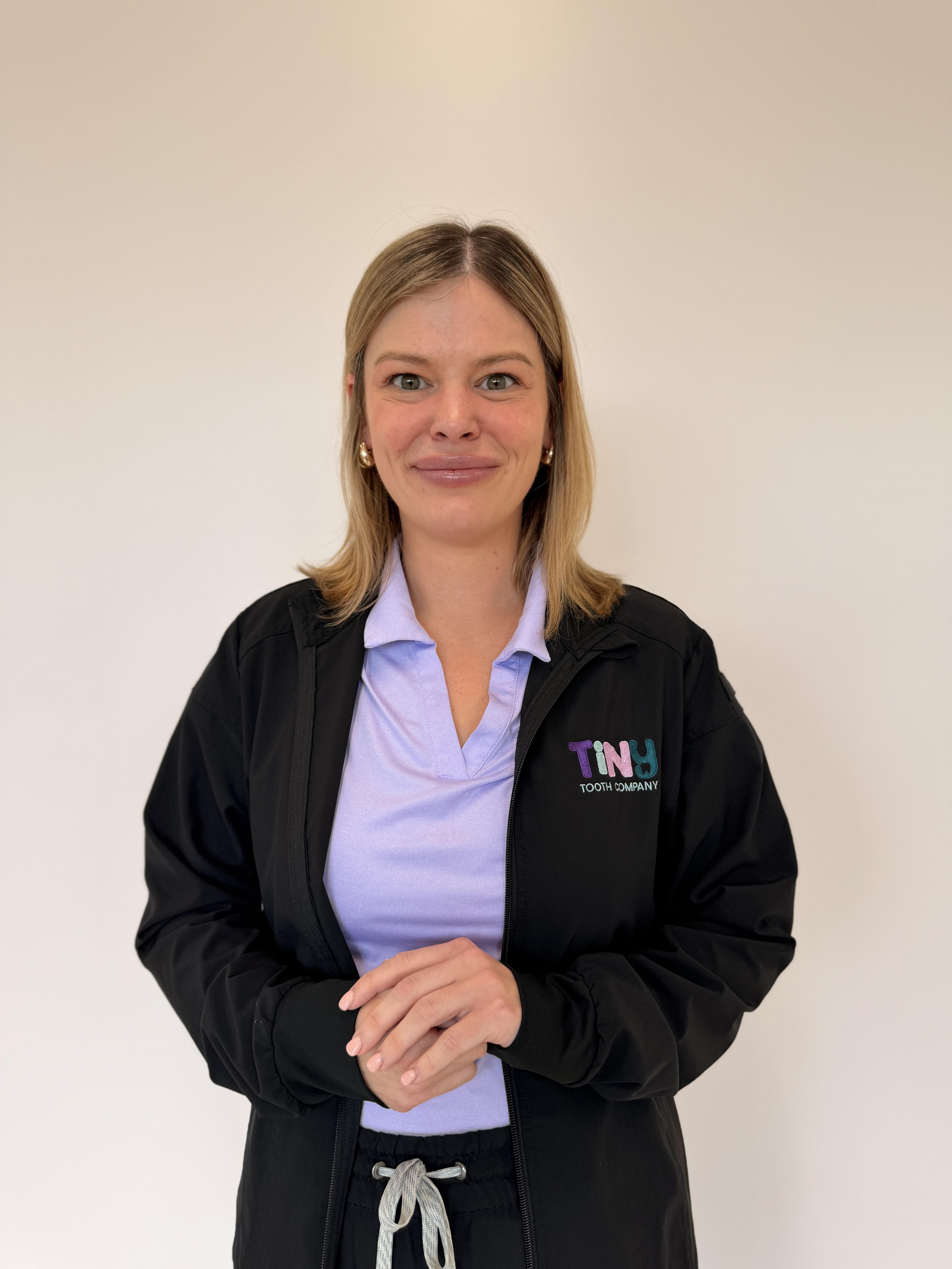 Madison, team member at Tiny Dental Company, smiling in a black Tiny Tooth Company jacket over a lavender scrub top