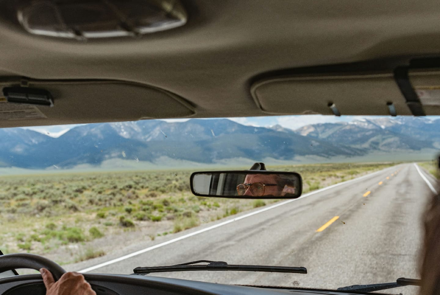 View from inside a vehicle of a driver wearing glasses seen in the rearview mirror driving along a deserted road with mountains in the distance.
