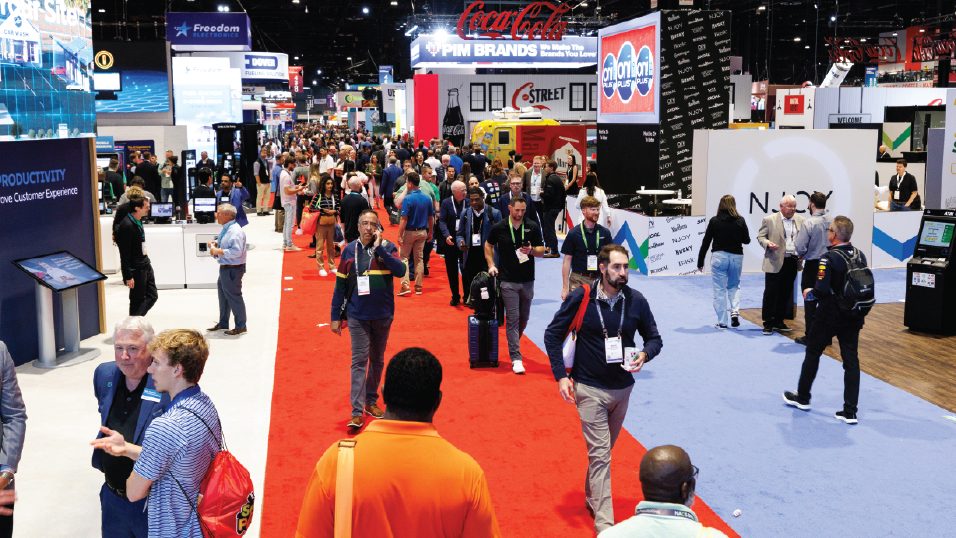 Attendees walking the main aisle of the NACS Show exhibit hall between booths.