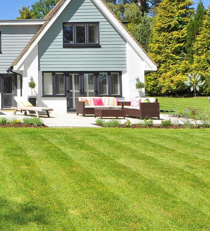 Modern house with a neatly cut green lawn, outdoor seating area with colorful cushions, and surrounded by trees.