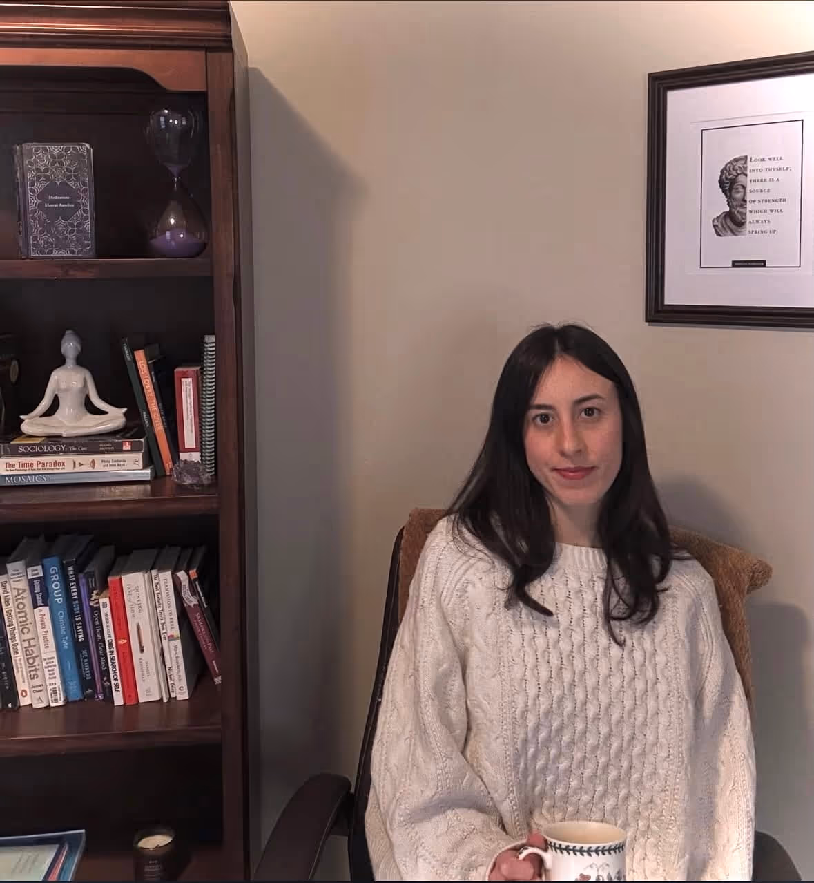 Savannah in a white sweater sitting and holding a patterned mug near bookcase and framed art.