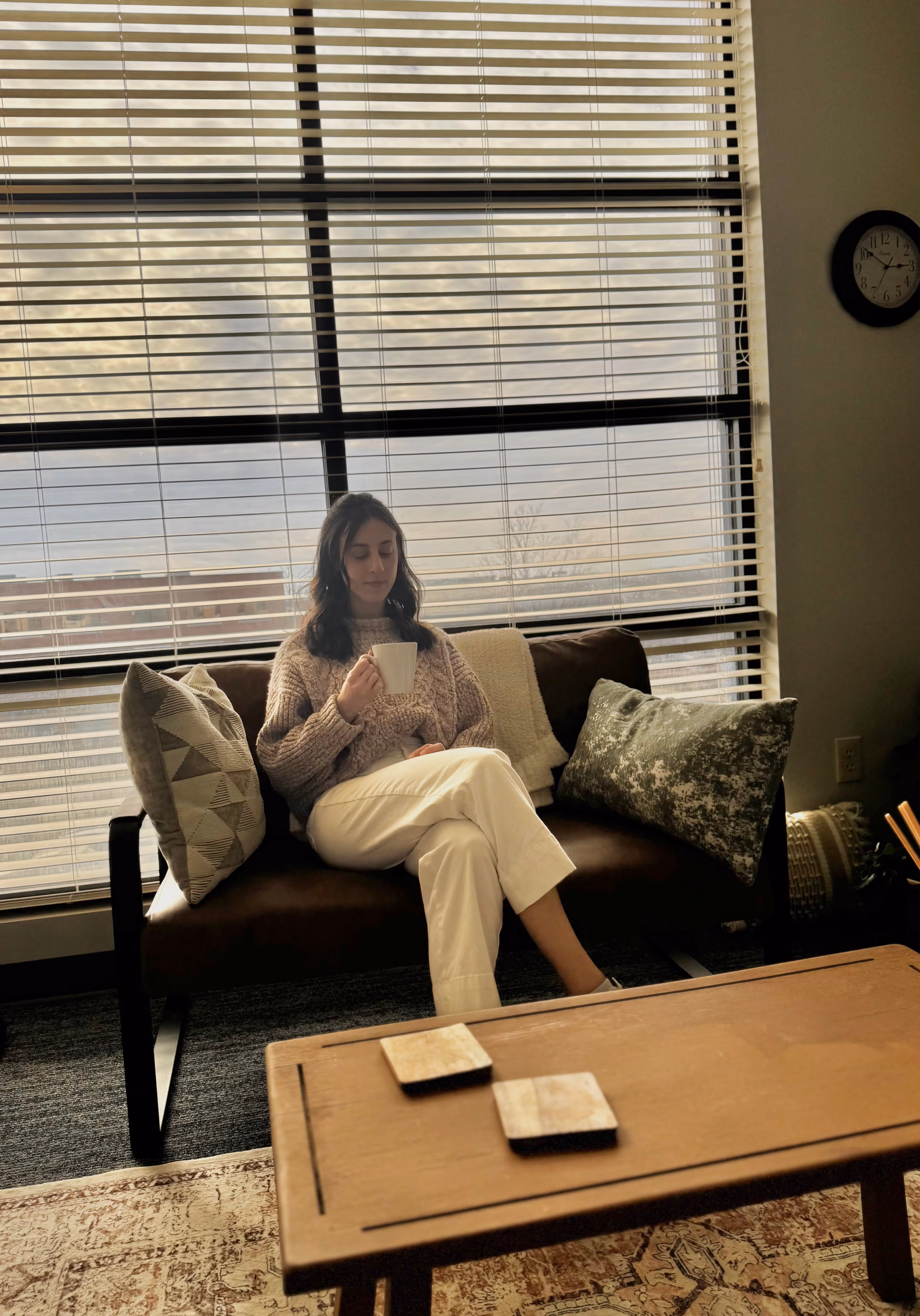 Savannah from Balance You Counseling Wellness, Therapy in Lexington, KY in white pants and beige sweater sitting on couch holding a mug in front of large window blinds. Therapist Lexington, KY