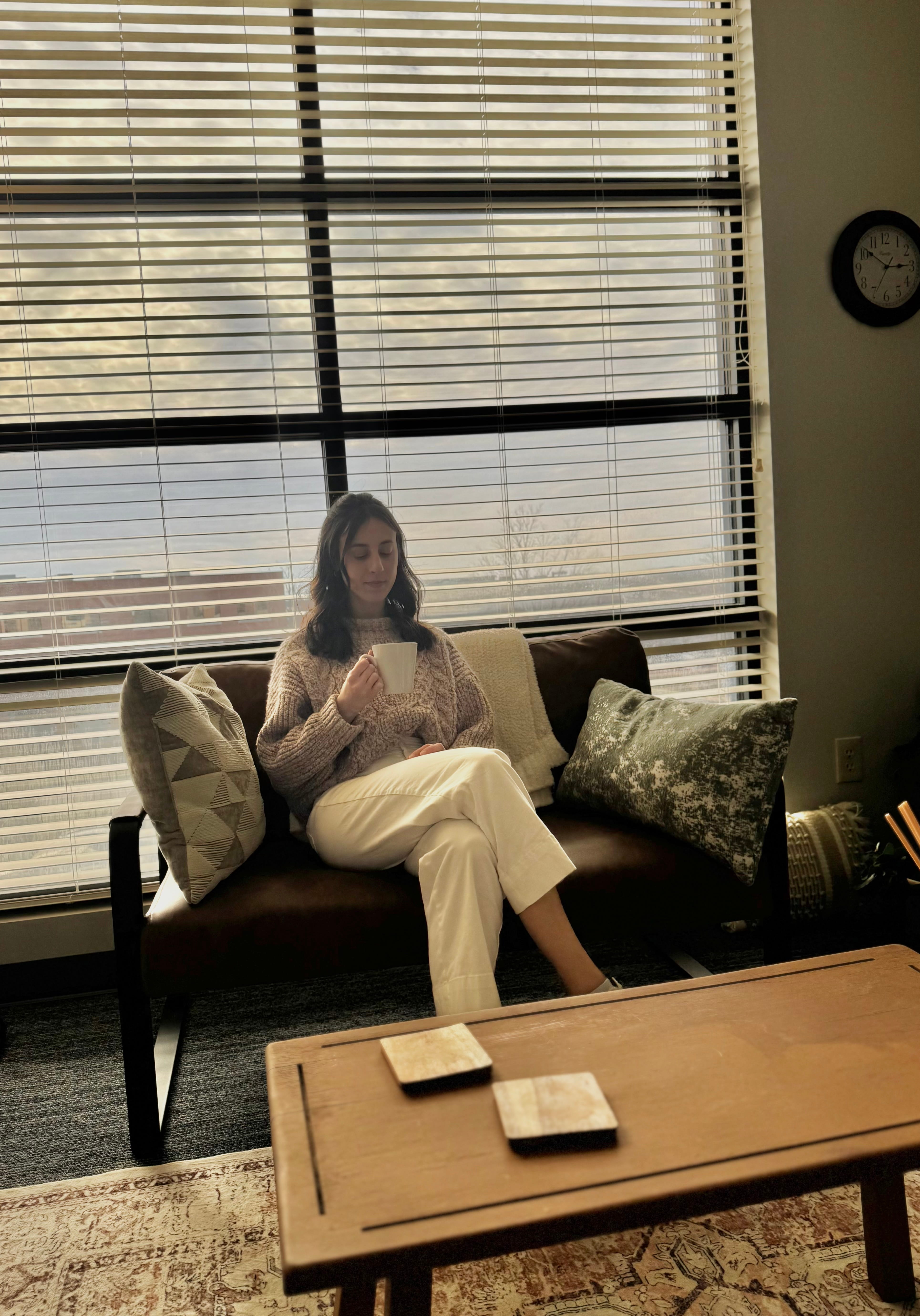 Savannah in white pants and beige sweater sitting on couch holding a mug in front of large window blinds. Therapist Lexington, KY