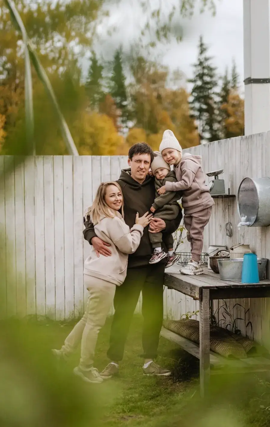 A young family outside in their back yard smiling at the camera, the small child standing on an outdoor kitchen bench