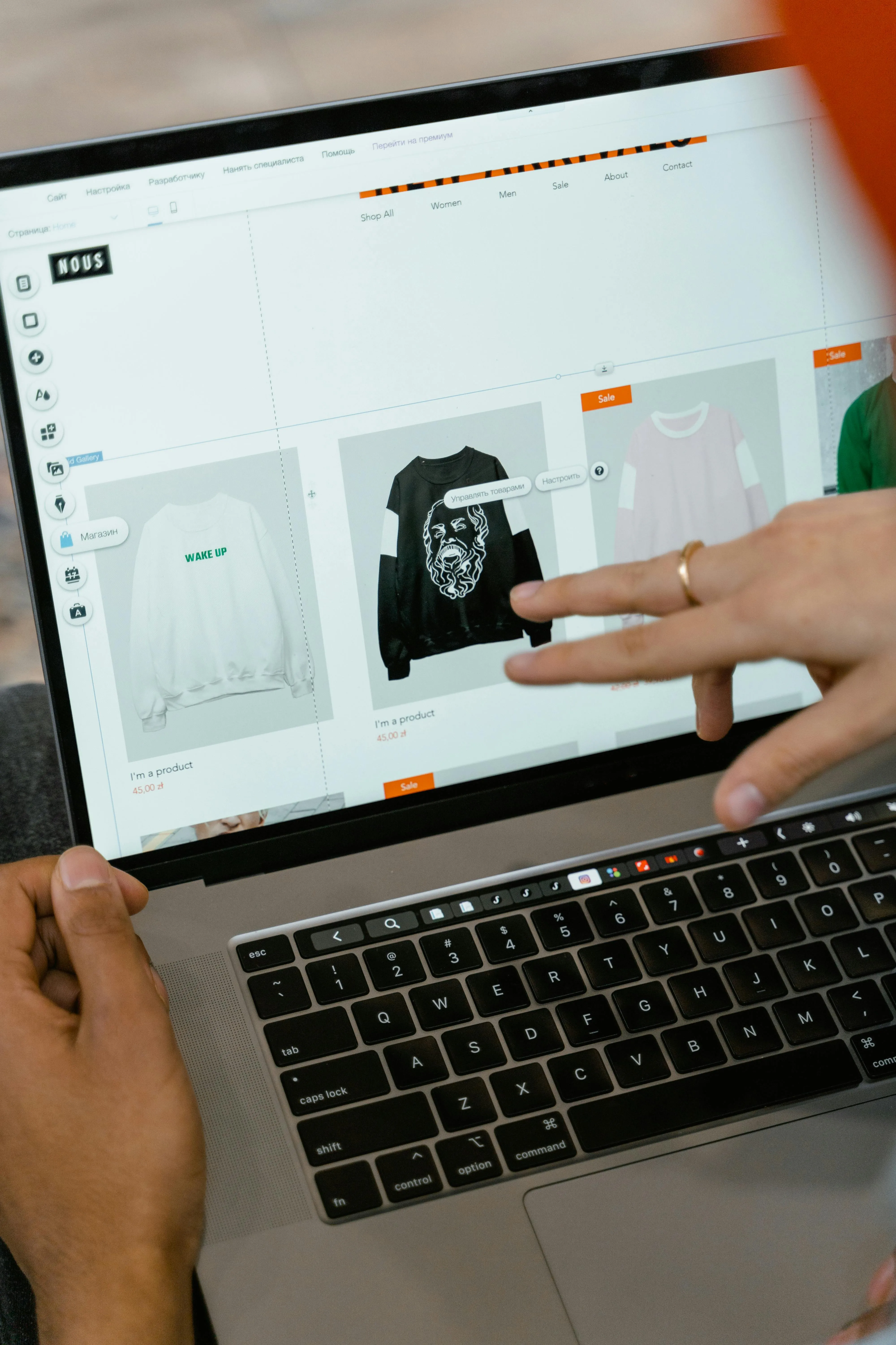 Person browsing an online store on a laptop, pointing at black sweatshirt with a face design.