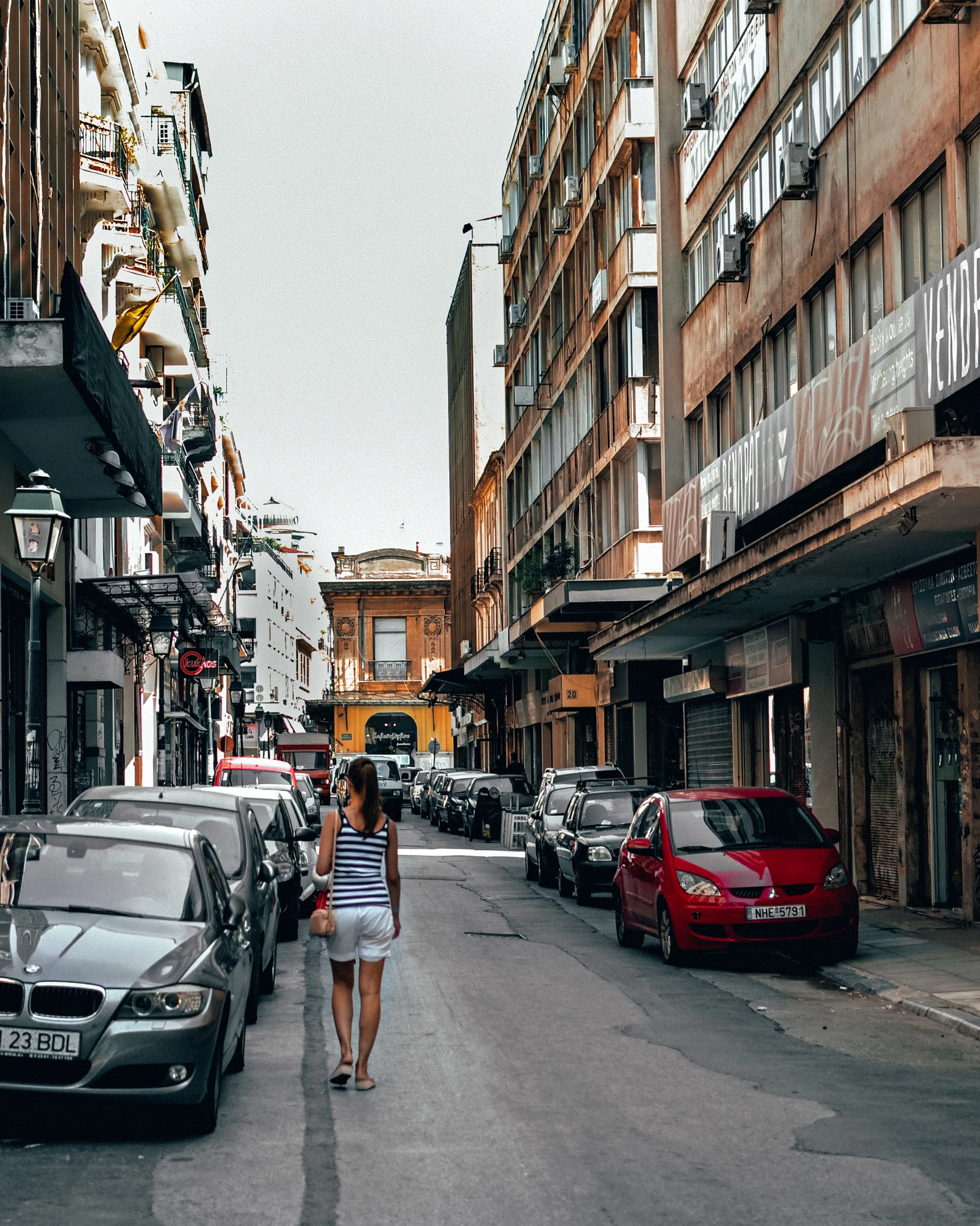 Woman in striped top walks down a narrow city street lined with parked cars and buildings.
