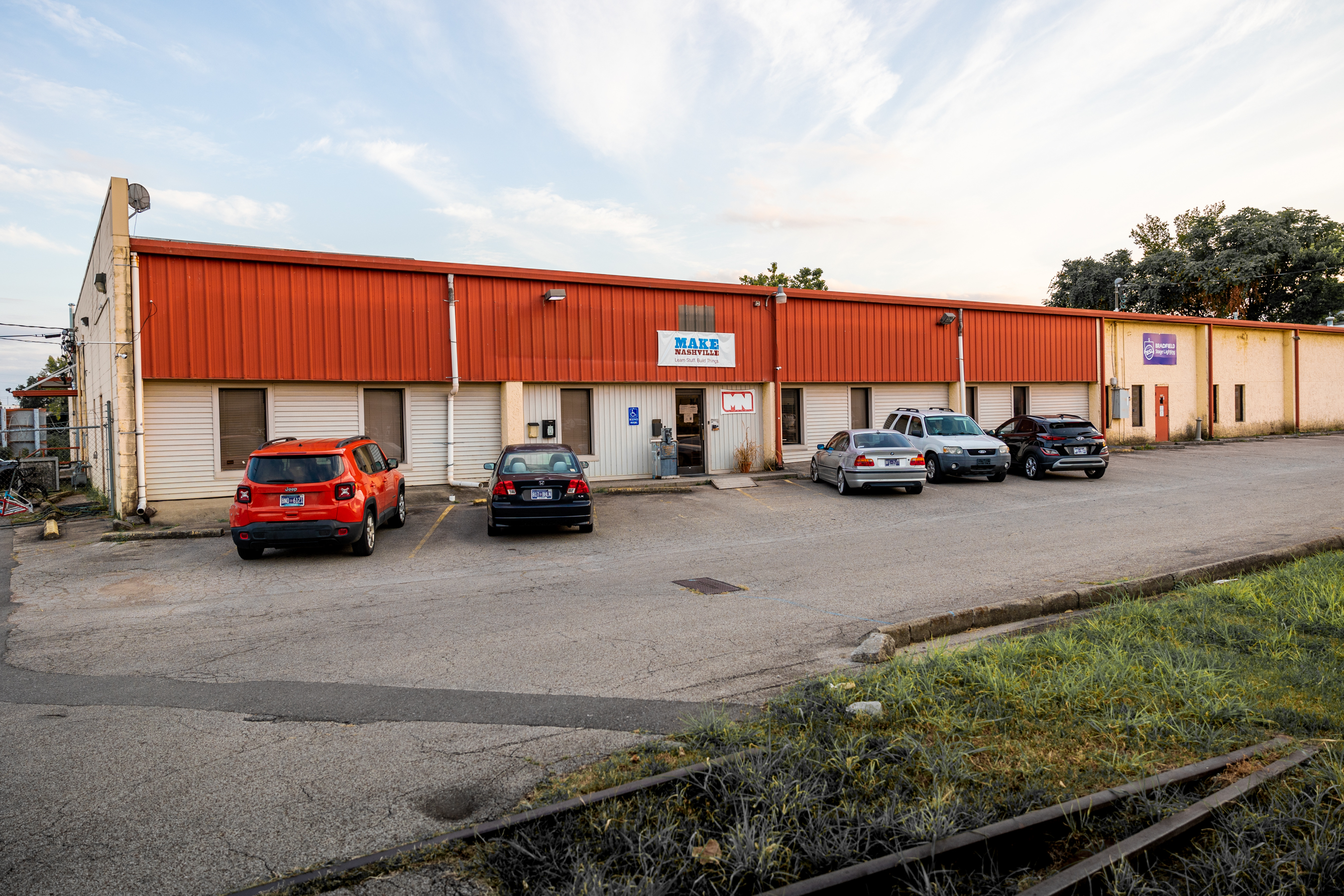Single-story commercial building with a red metal roof, several parked cars in front, and signs for Make Nashville.