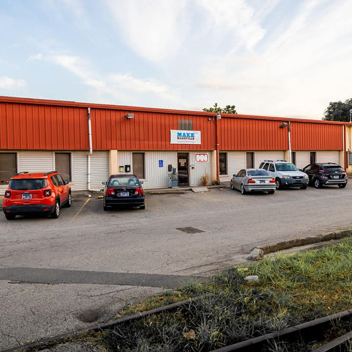 Single-story commercial building with a red metal roof, several parked cars in front, and signs for Make Nashville.