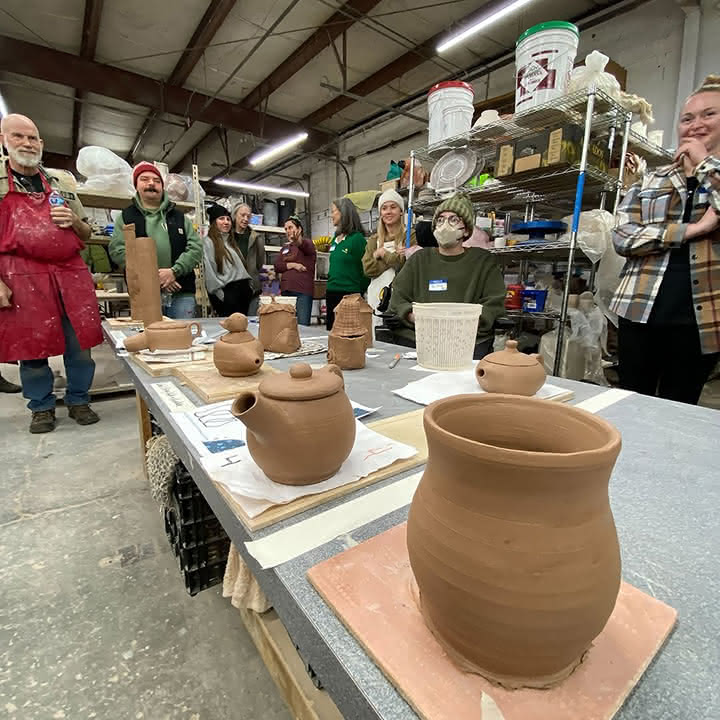 Group of people standing and sitting around a table displaying various handmade pottery pieces in a workshop.